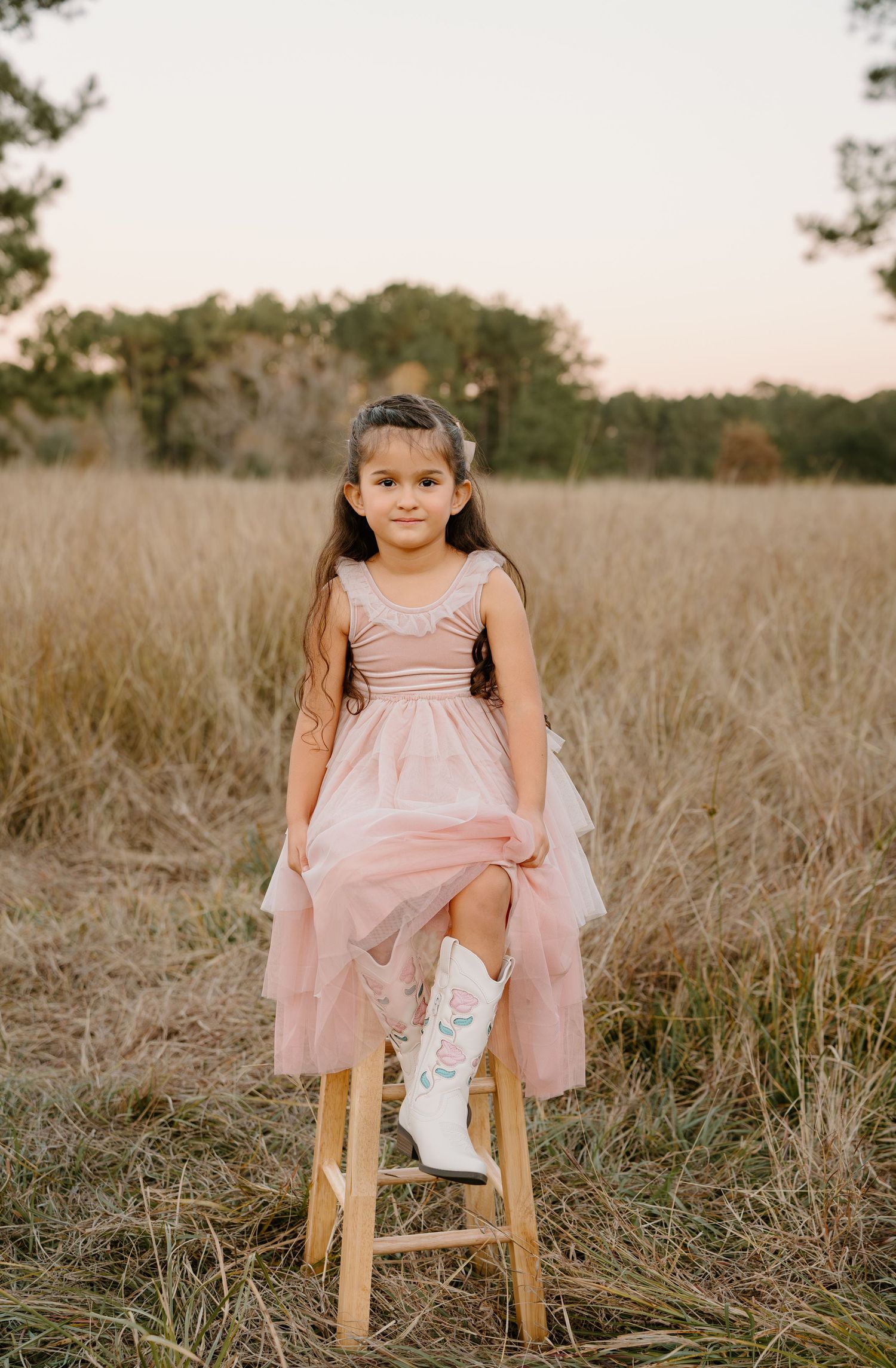 Young girl in pink tulle dress and white cowboy boots sits on wooden stool in golden field at sunset.