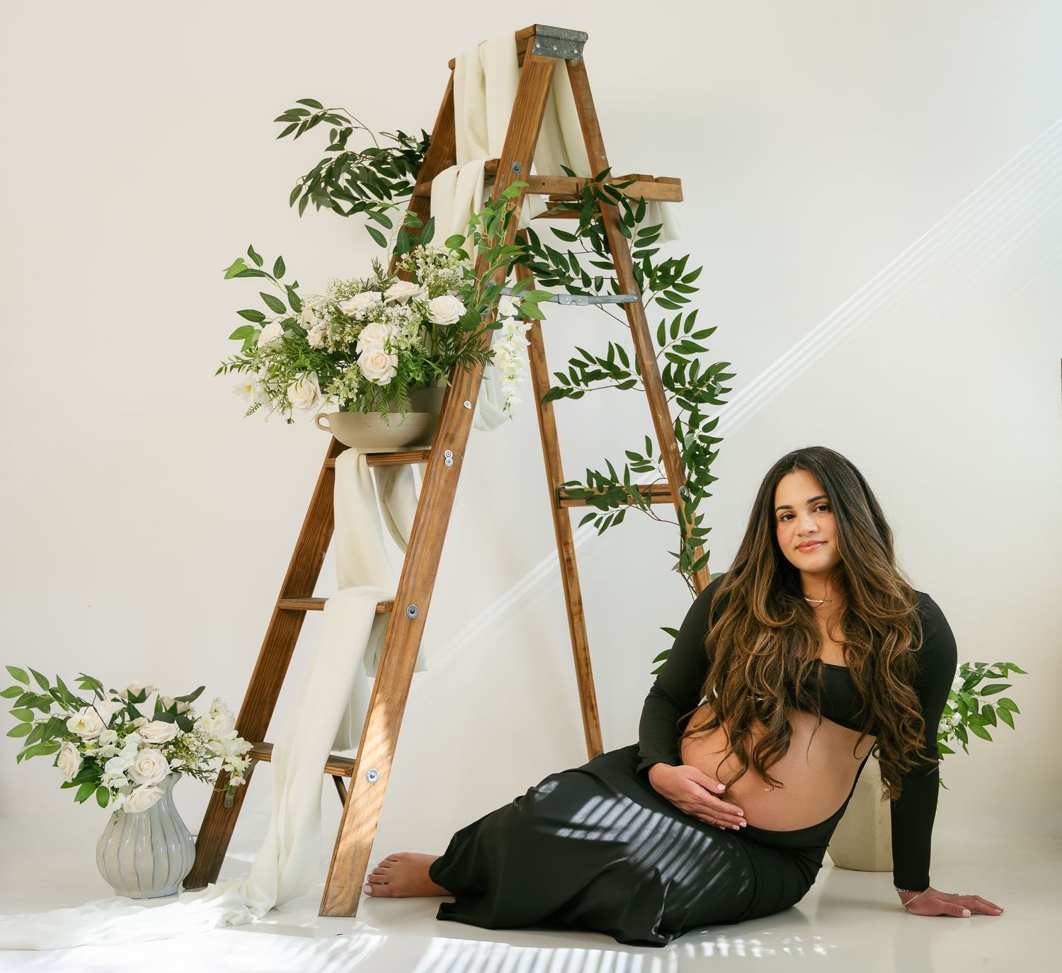 Pregnant woman in black outfit sitting on floor near a wooden ladder decorated with white flowers.