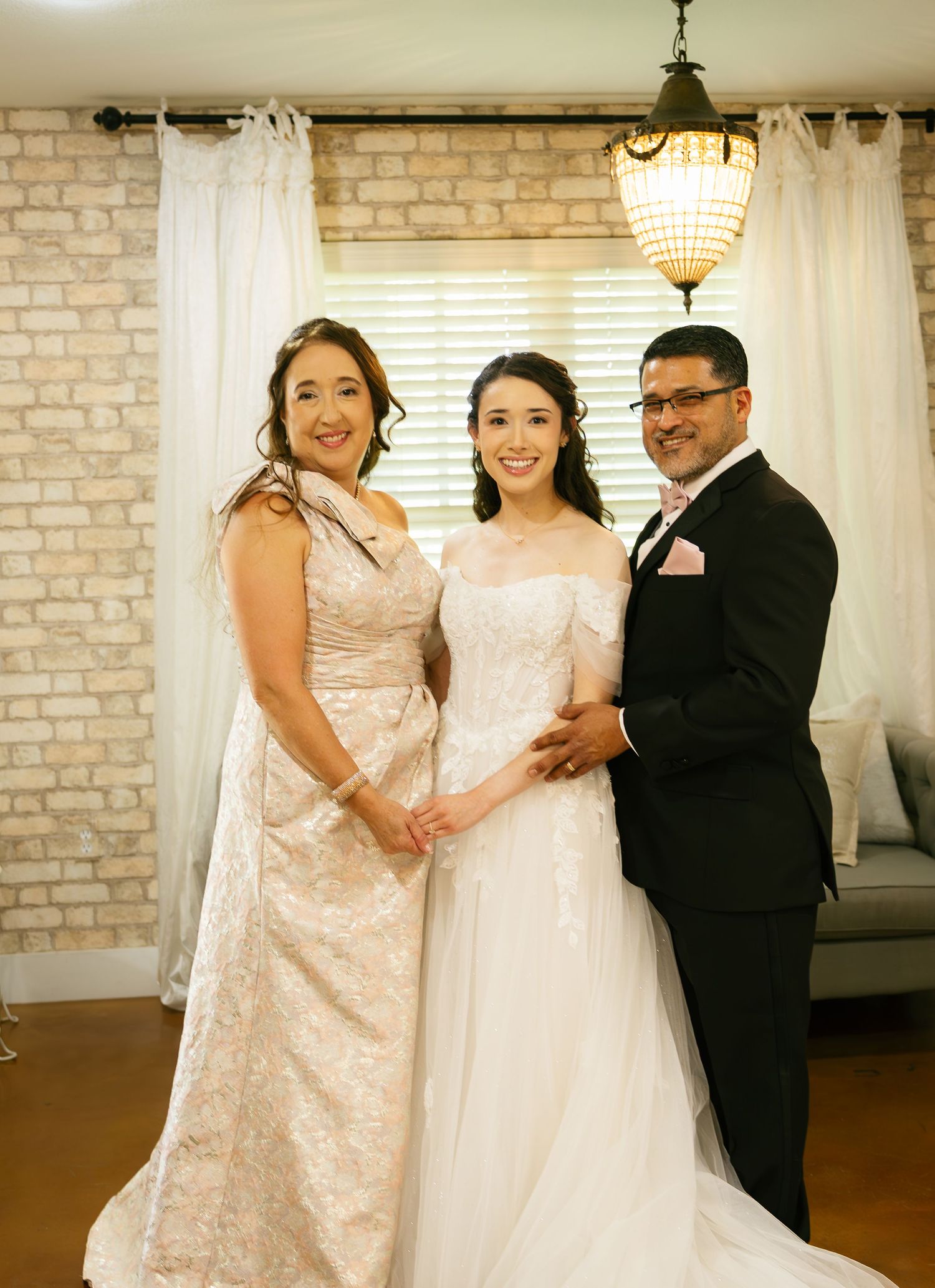 A smiling bride in a white lace gown poses with her parents in formal attire inside an elegant venue.