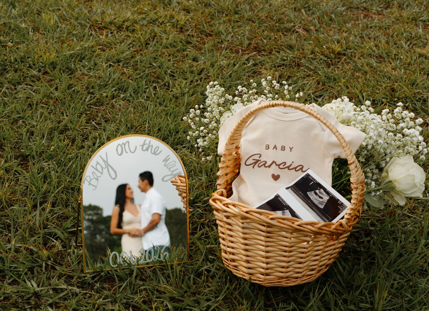 Pregnancy announcement basket with baby onesie, ultrasound photos, white flowers, and a personalized couple photo sign.