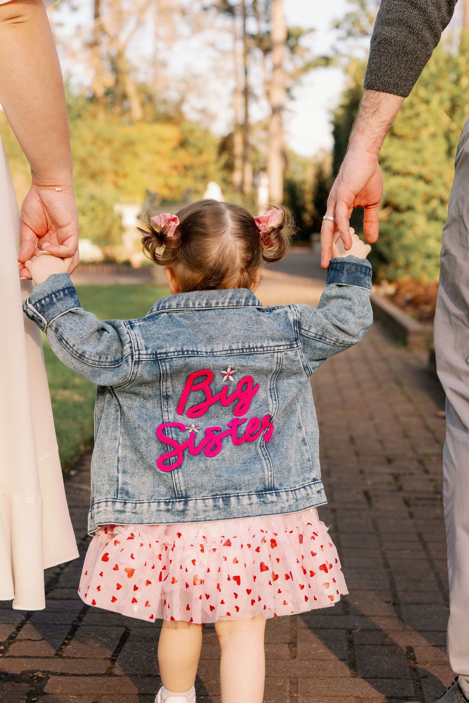 Toddler girl wearing a denim jacket with pink Big Sister patch holds parents hands on a brick path.