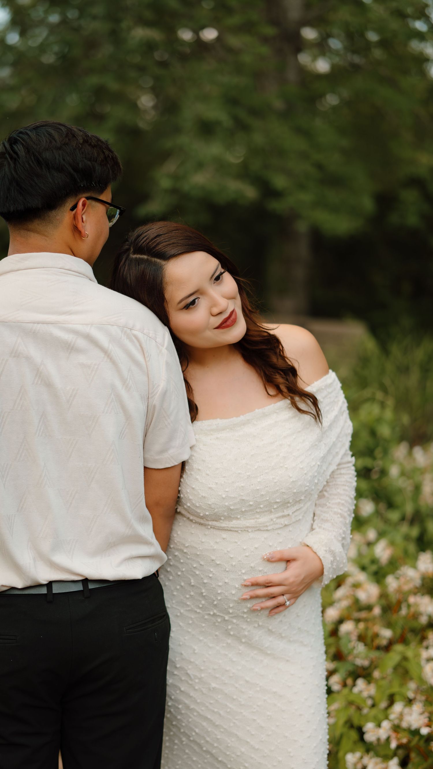 A pregnant woman in a white off-shoulder gown embraces her partner outdoors among blooming flowers and lush greenery.