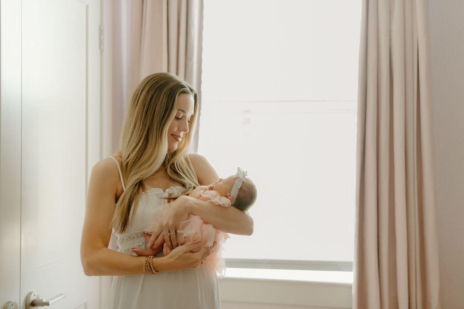 A smiling mother in a white dress holds her newborn baby near a bright window with soft pink curtains.