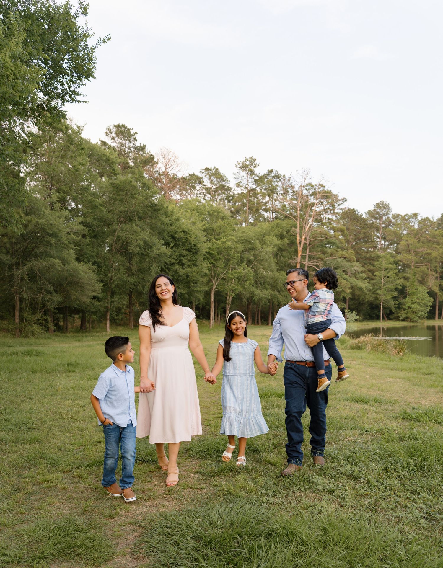 Family of five walking outdoors in a green park, parents holding children's hands, smiling together on a sunny day.
