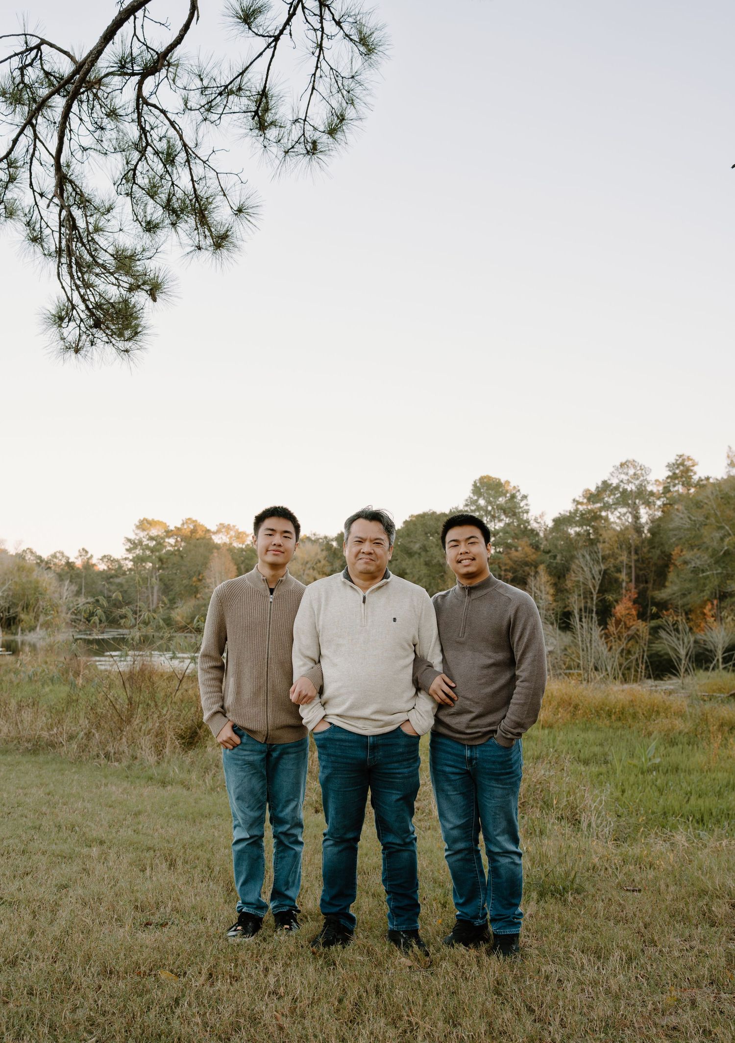 Three men standing together in a grassy field with trees and a pond in the background during golden hour.