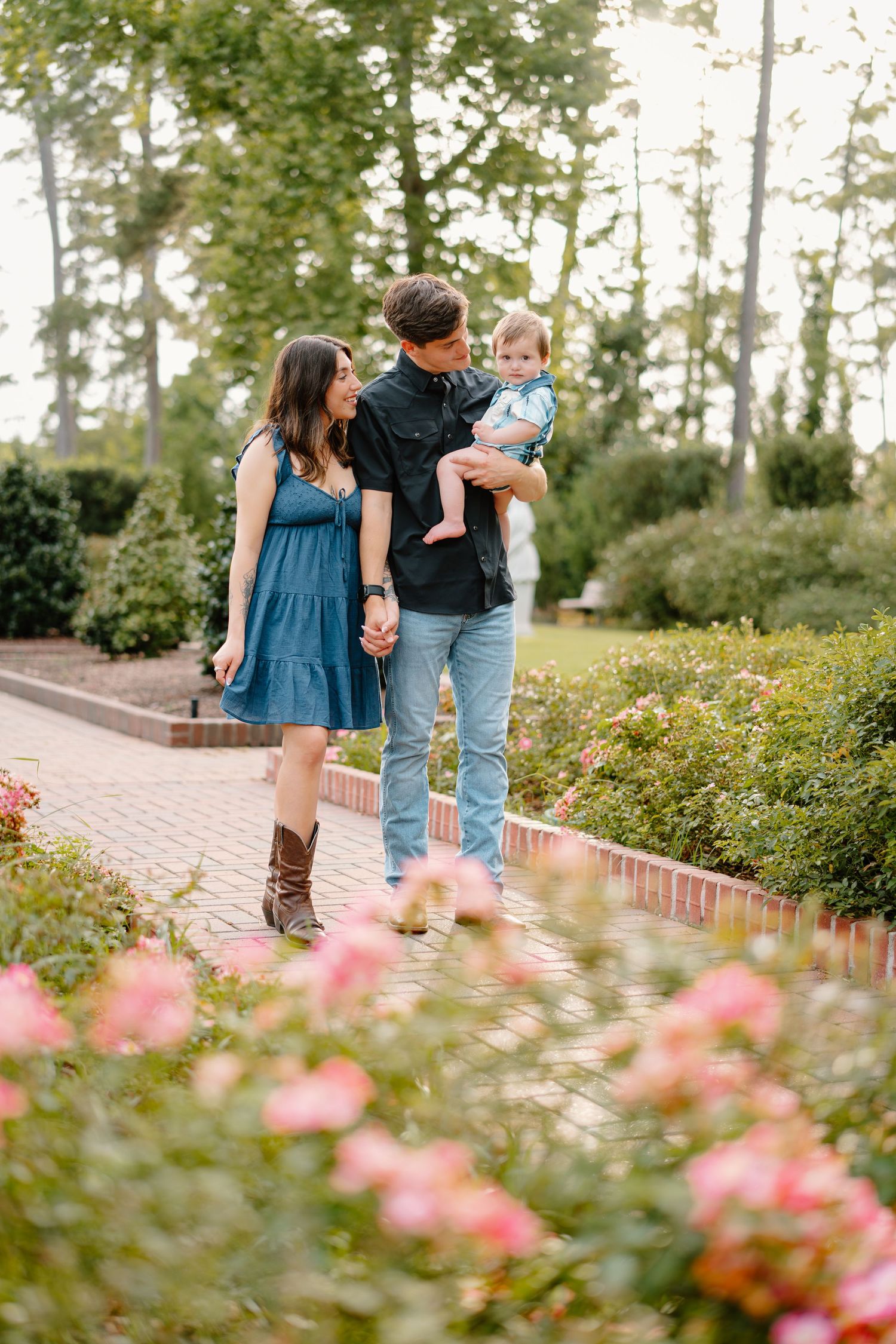 A young family poses on a brick garden path surrounded by pink roses and lush greenery, parents holding their toddler.