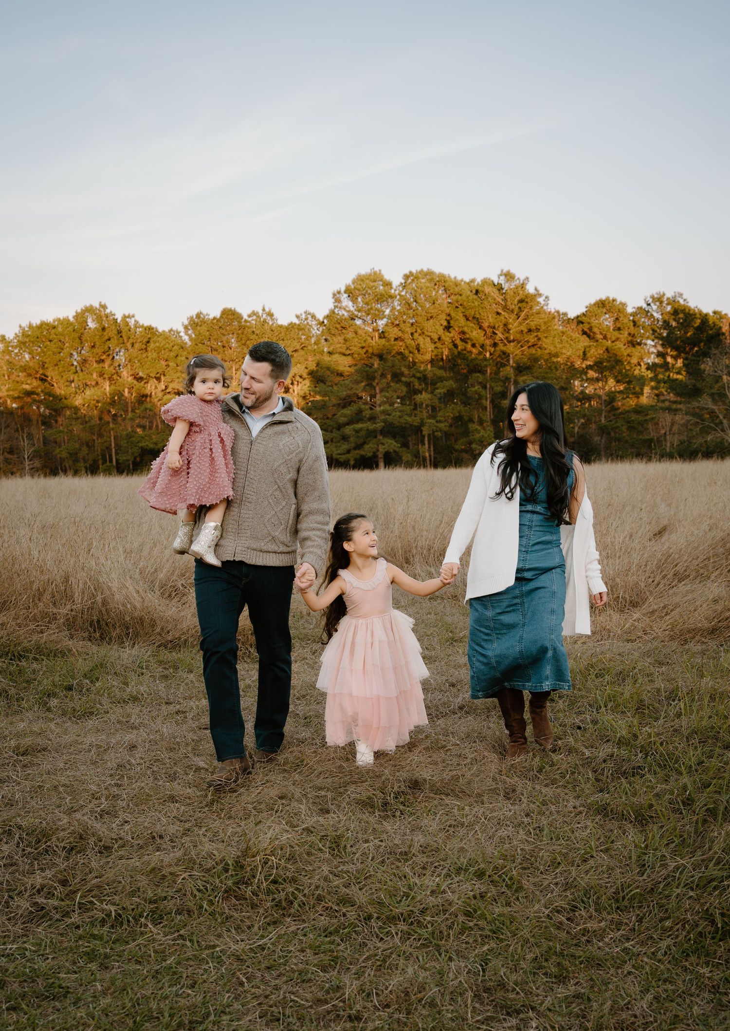 Family of four walking in golden field at sunset, parents holding hands with young girls in pink dresses.