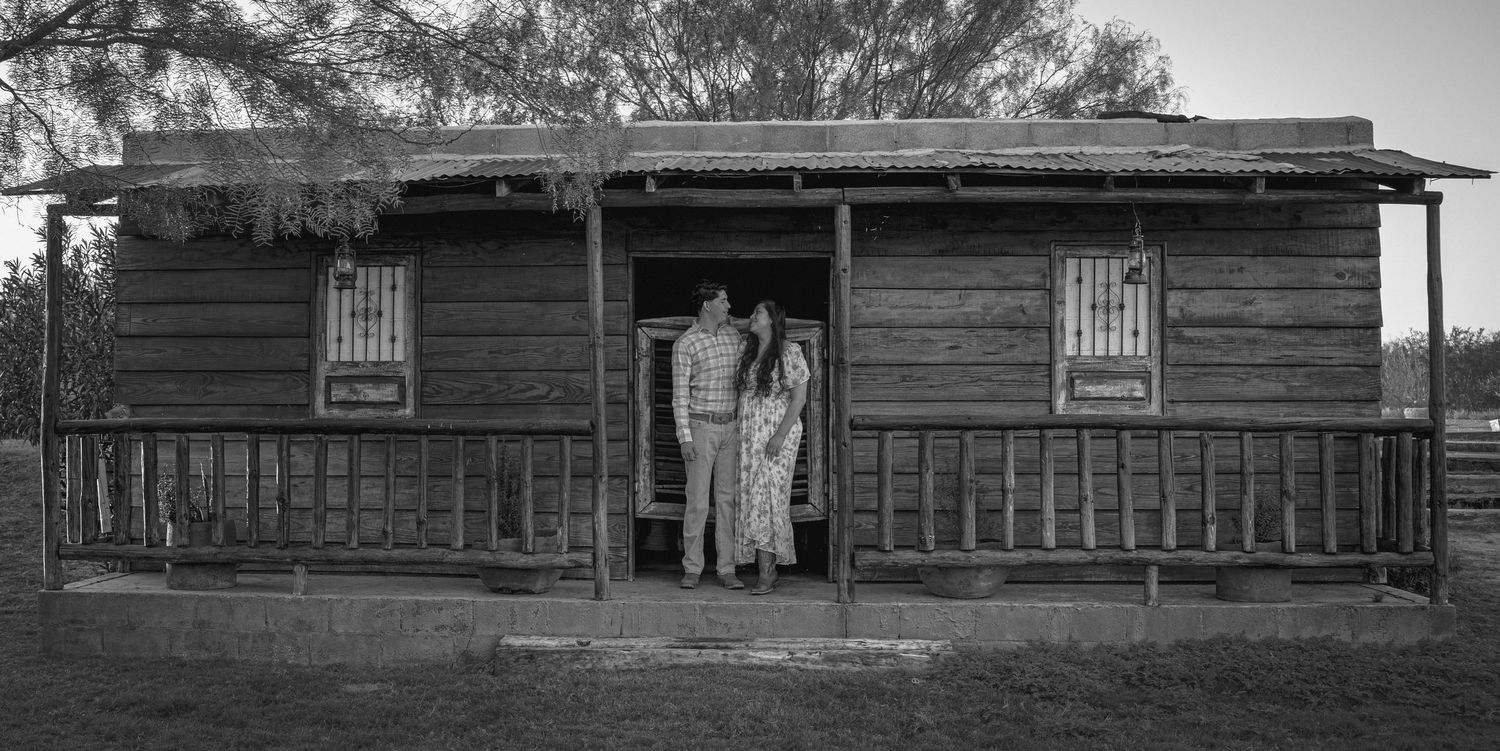 Black and white photo of couple standing in doorway of historic rustic log cabin with covered porch.
