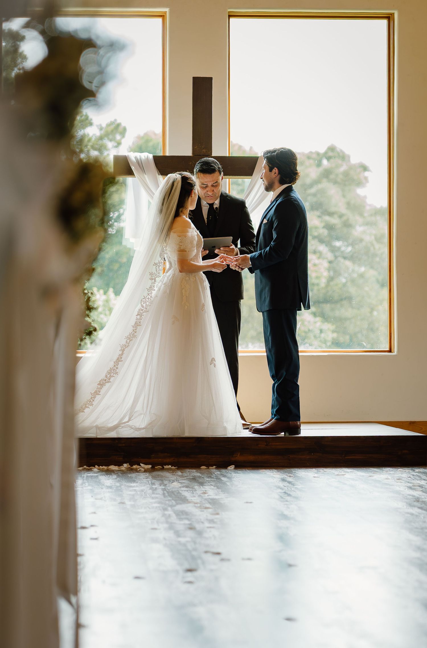 Bride and groom exchanging rings at altar before officiant in sunlit chapel with wooden cross backdrop.