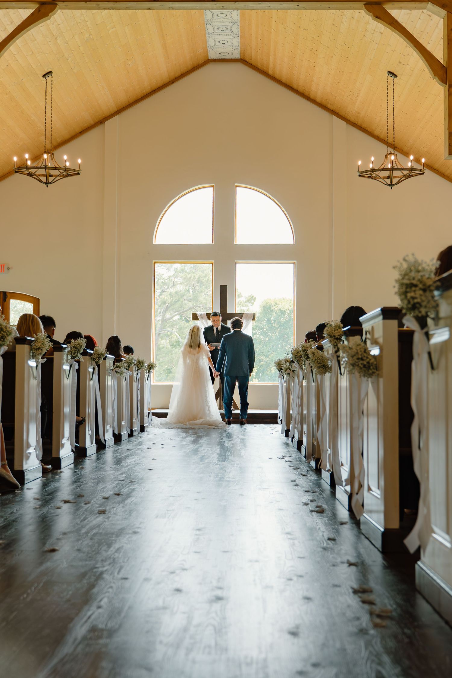 A bride and groom stand at the altar of a charming white chapel with wooden ceiling and floral-adorned pews.