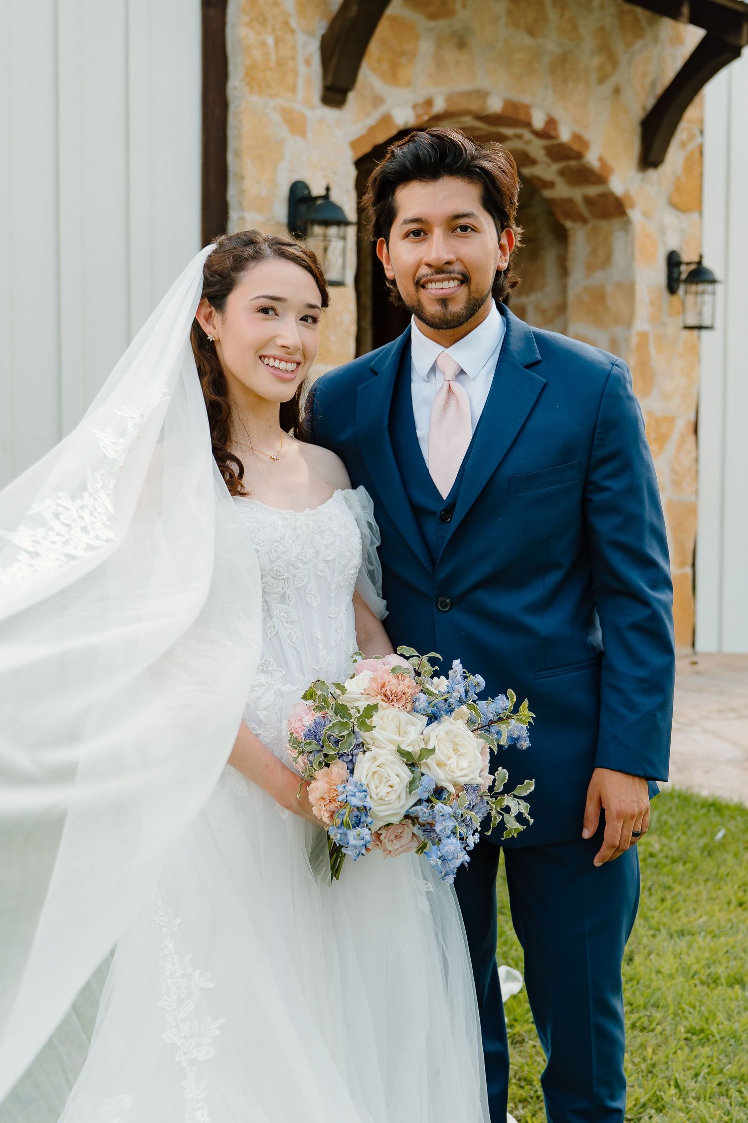 Smiling bride and groom posing outdoors holding a blue and white floral bouquet.