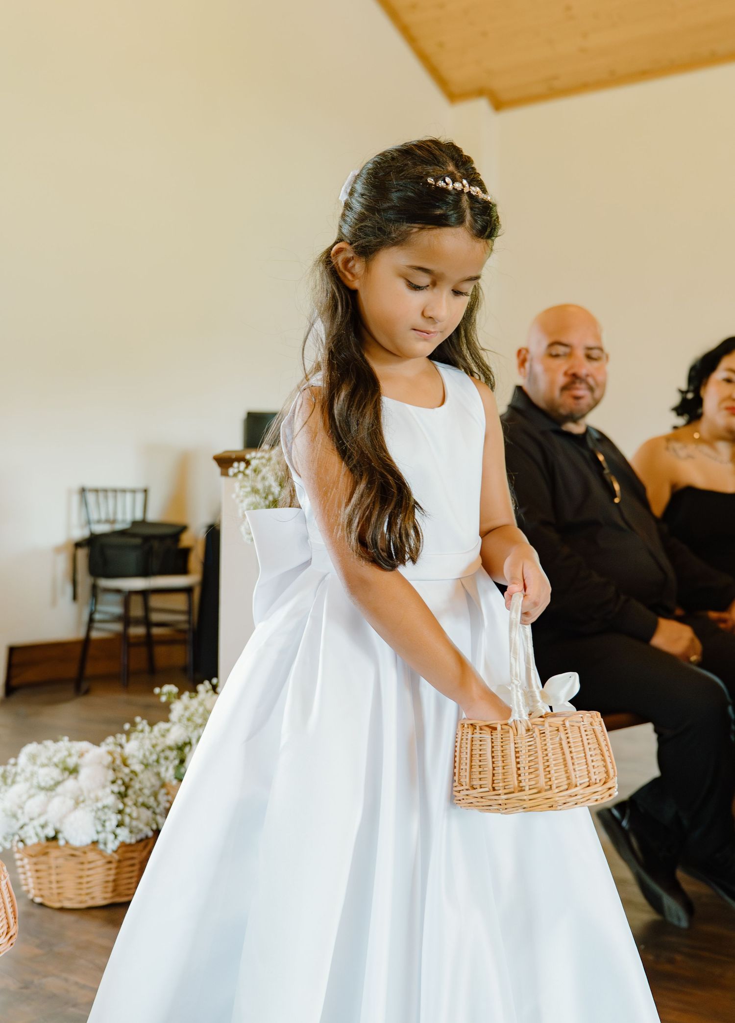 Young flower girl in a white dress carries a wicker basket during a wedding ceremony indoors.