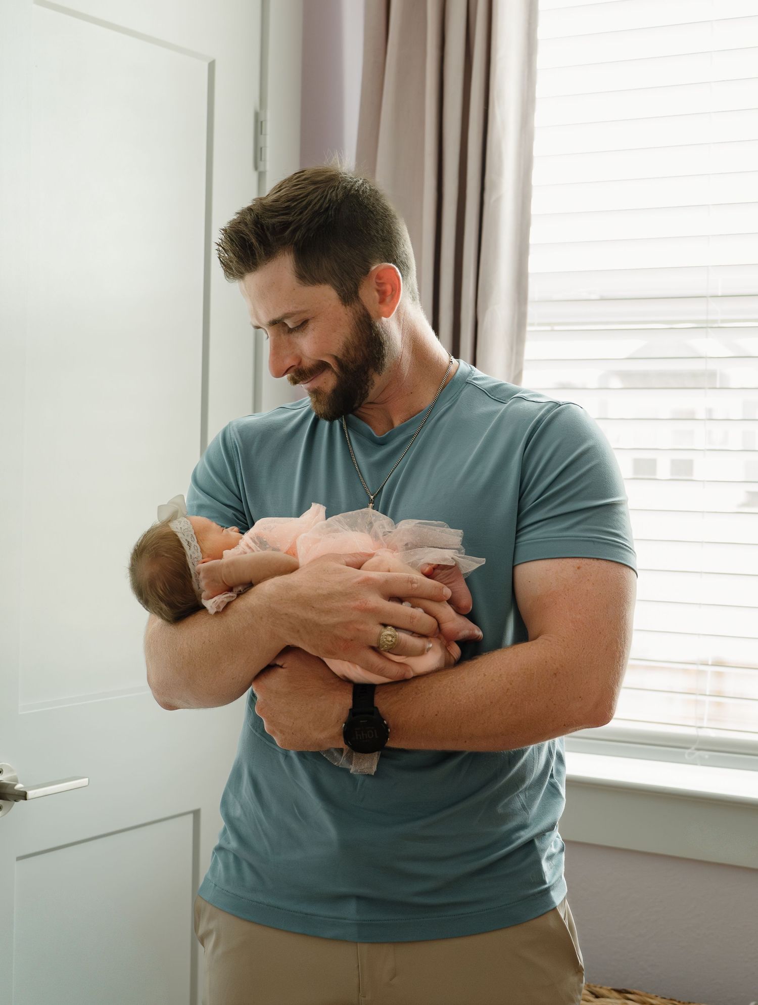 A muscular bearded man in a teal shirt tenderly holds a sleeping newborn baby near a bright window indoors.