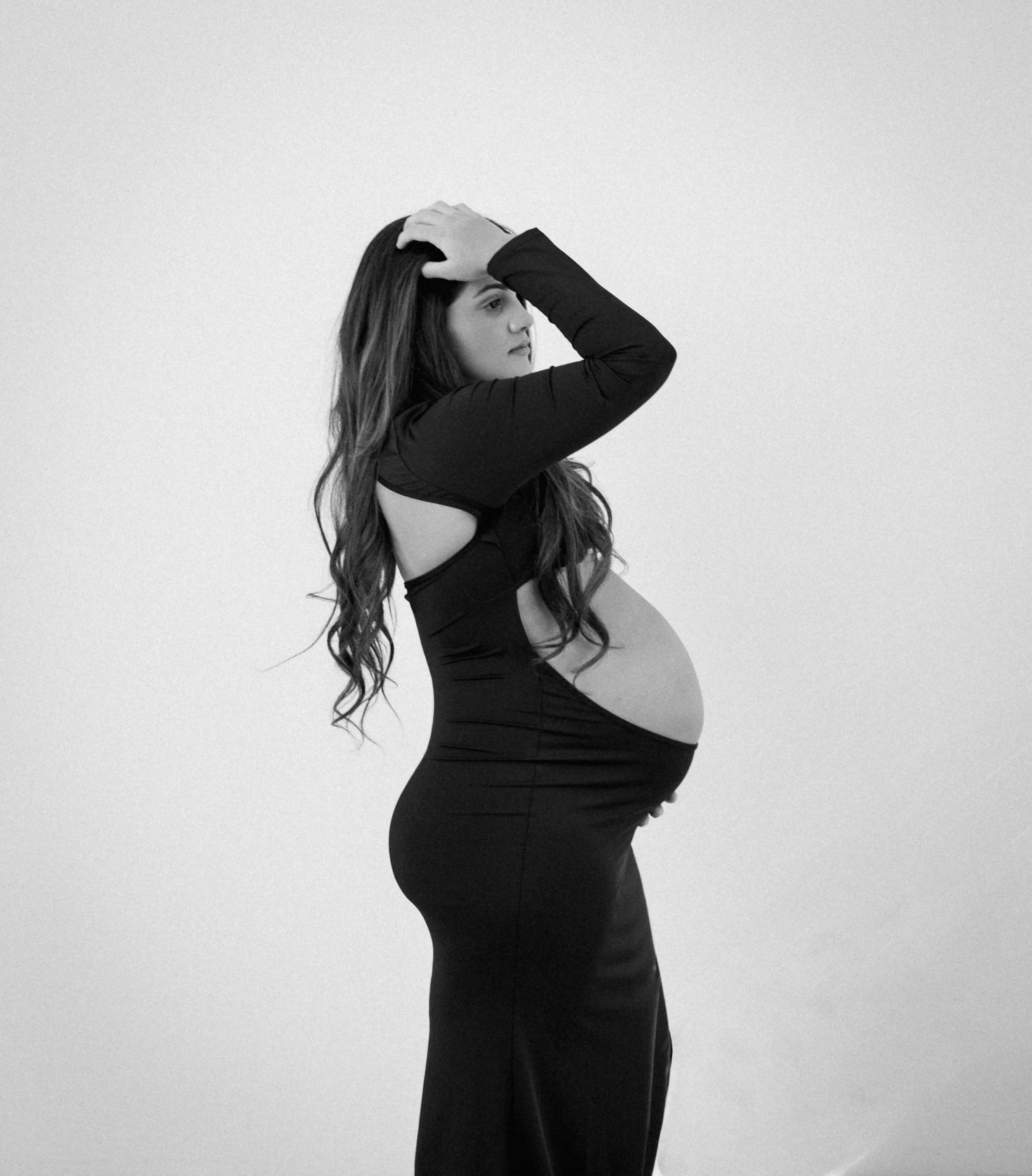 Black and white maternity photo of pregnant woman in backless black dress posing sideways.