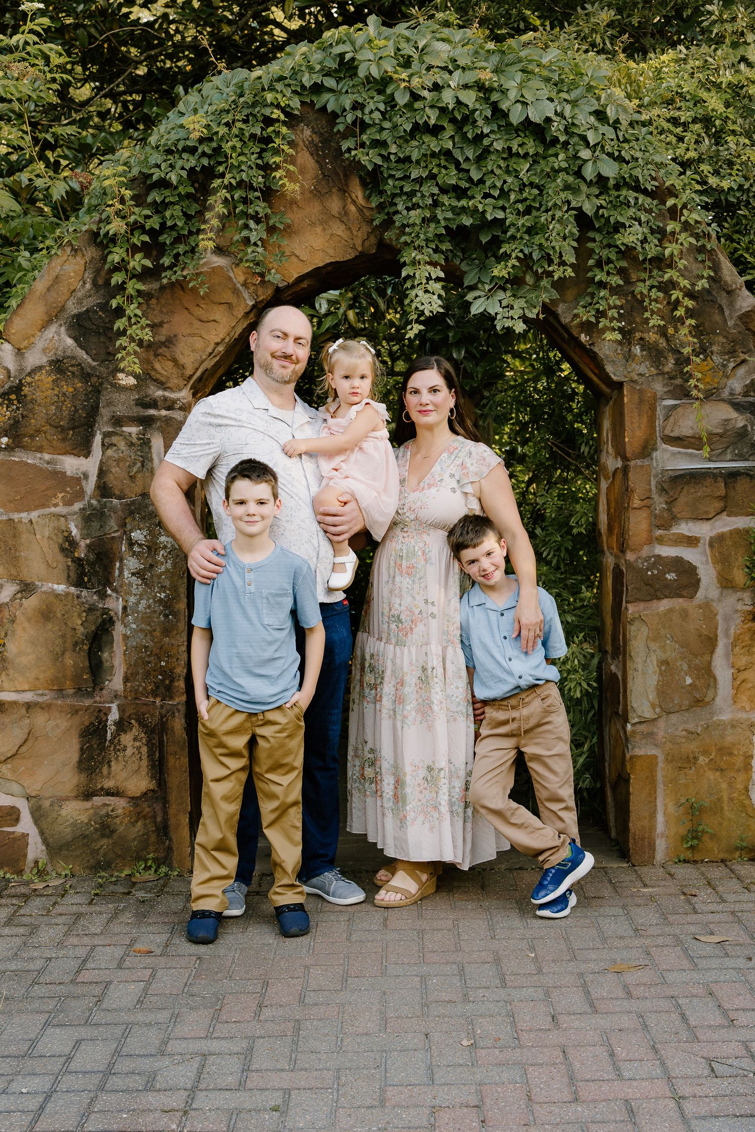 Family of five posing under a stone arch covered in greenery, smiling outdoors in a garden setting.