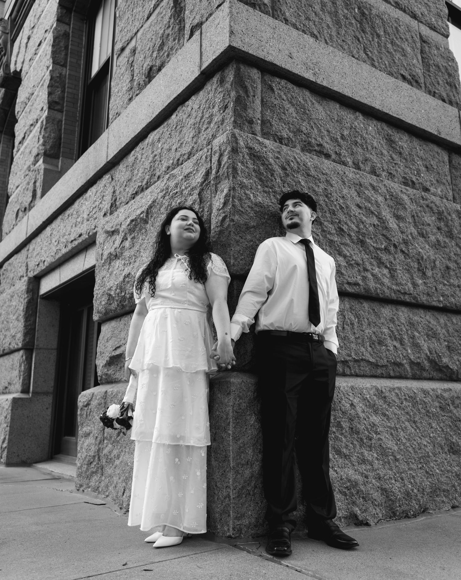 Black and white photo of bride and groom holding hands while leaning against historic granite building corner.