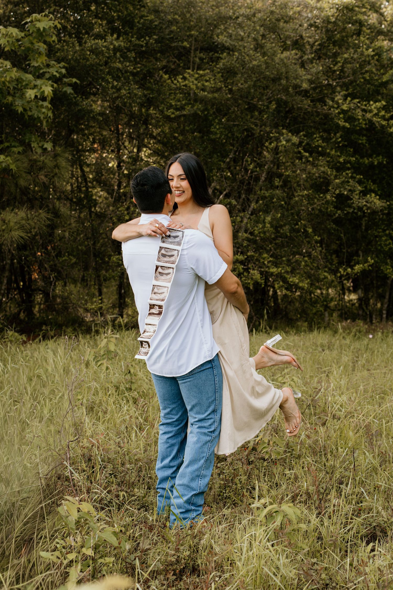 Man lifting smiling woman in beige dress outdoors, ultrasound photos tucked in back pocket for pregnancy announcement.