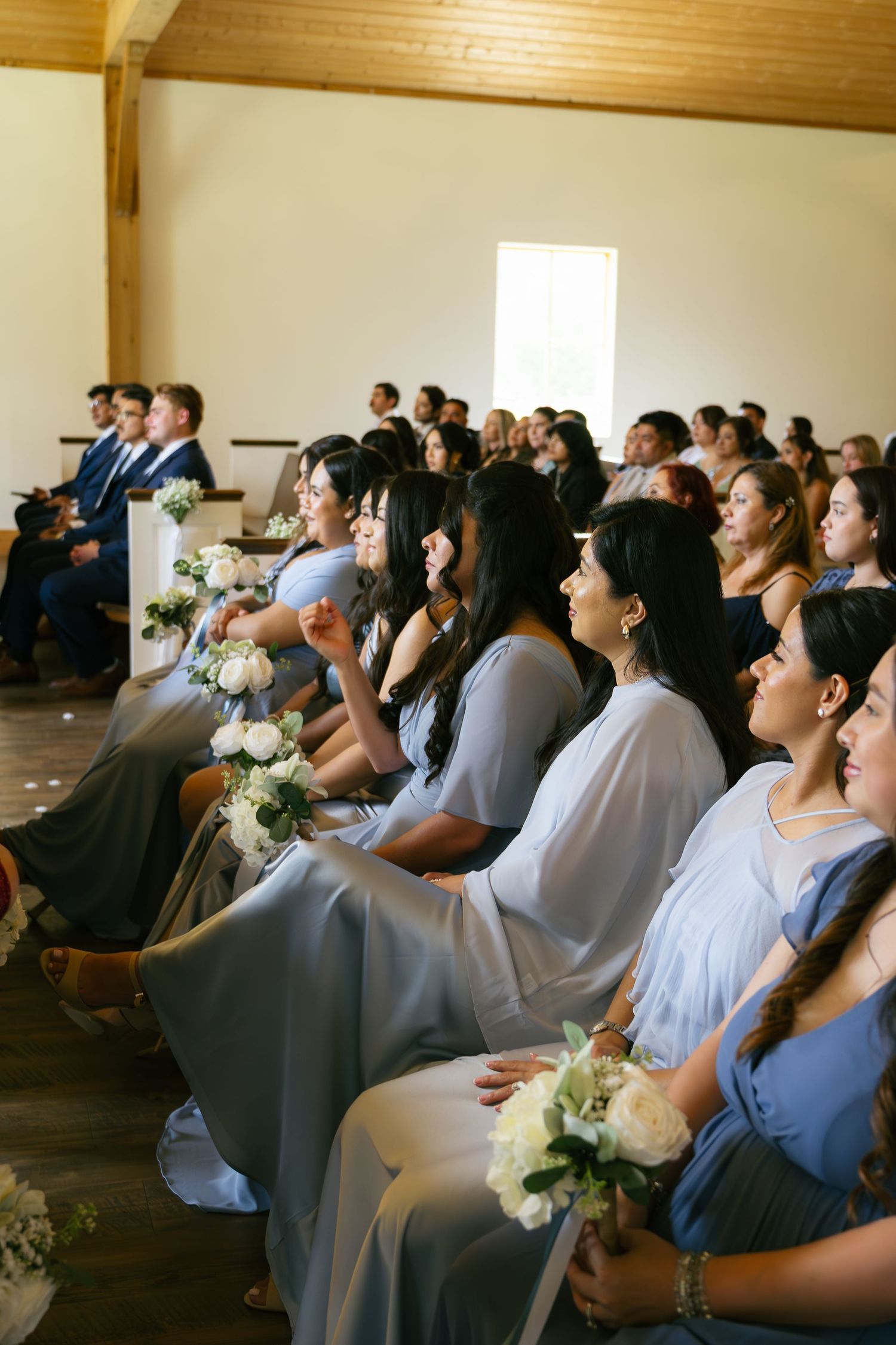 Bridesmaids in blue dresses holding white rose bouquets seated in church pews during wedding ceremony.