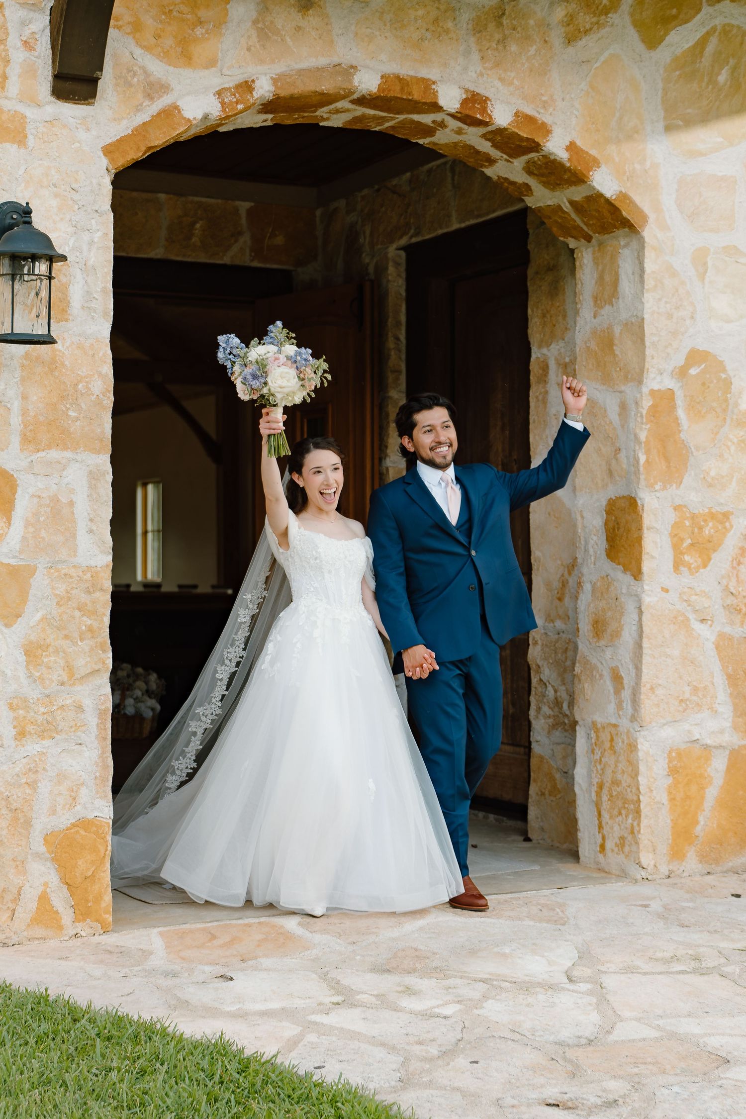 Joyful newlyweds exit stone arch venue, bride in white gown holding bouquet, groom in navy suit, both raising arms in celebration.