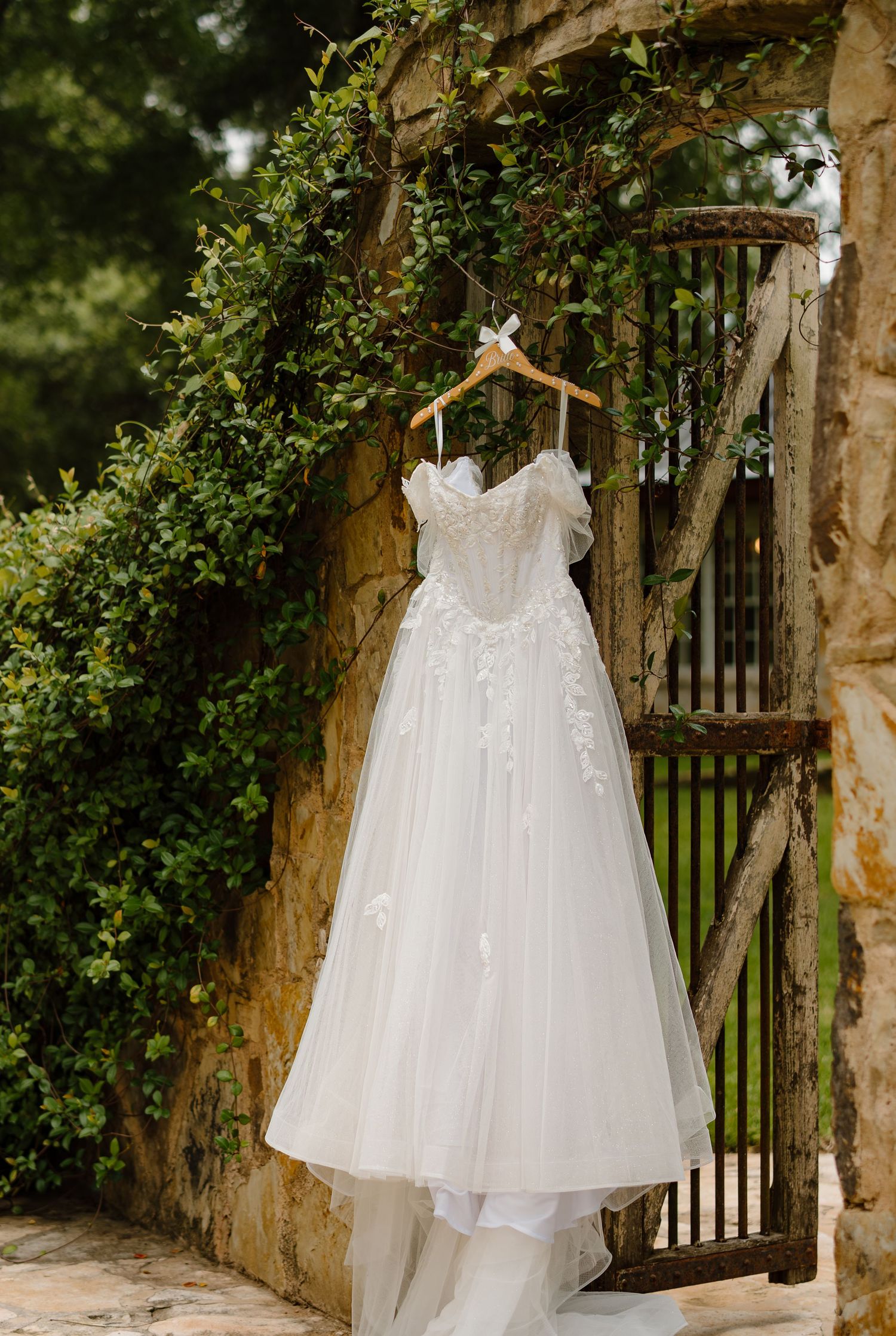 Elegant white lace A-line wedding dress hanging on wooden hanger against stone wall with lush greenery backdrop.