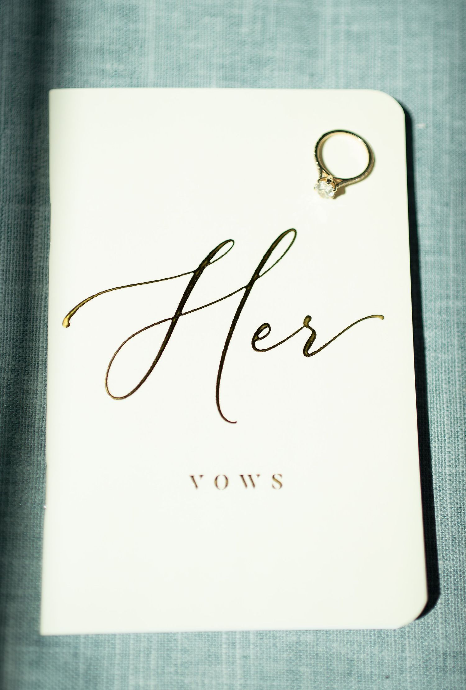 White vow book with gold script reading Her Vows, topped with a diamond engagement ring on blue fabric.