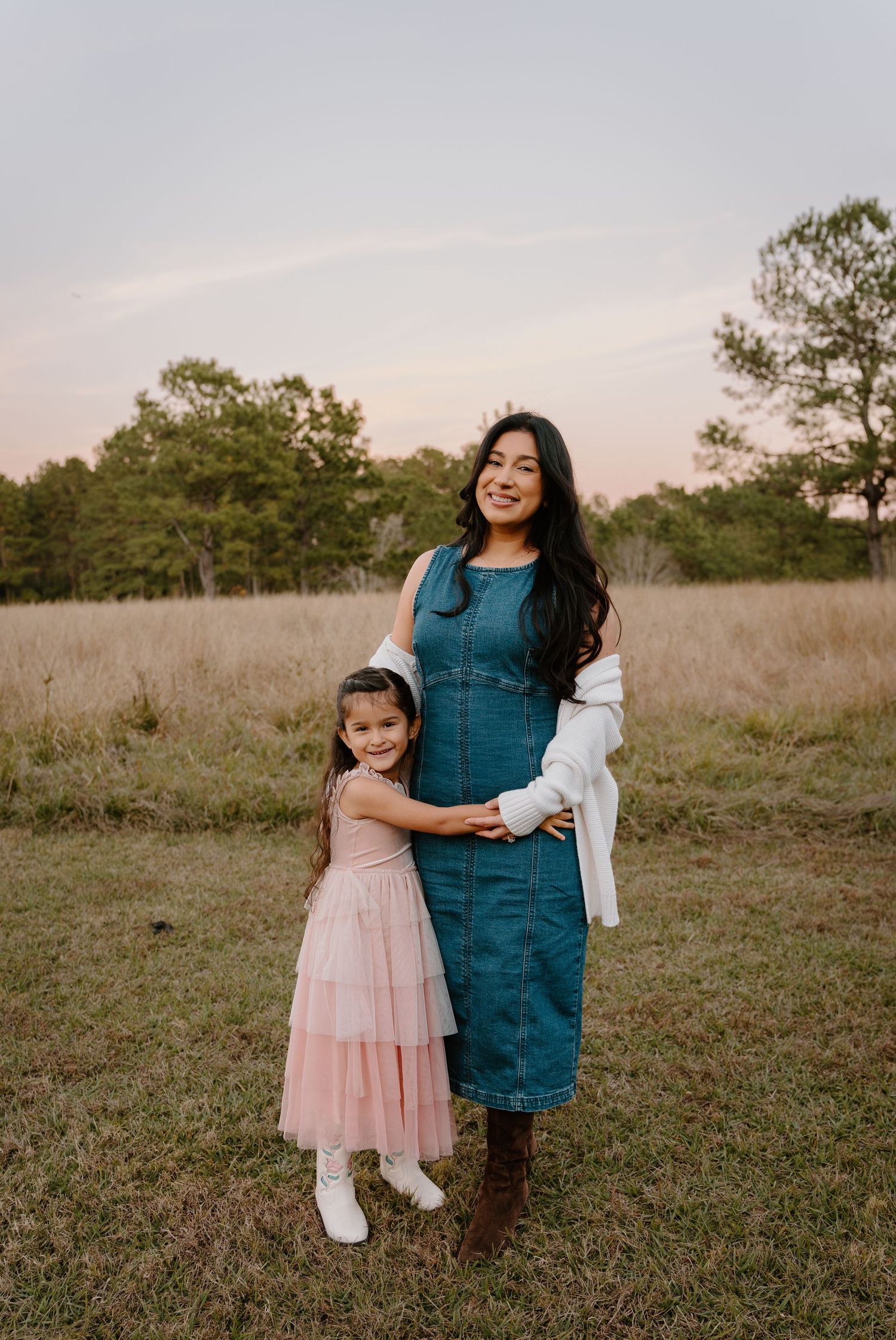 A woman in a teal denim dress and a young girl in a pink dress pose together in a golden field at sunset.