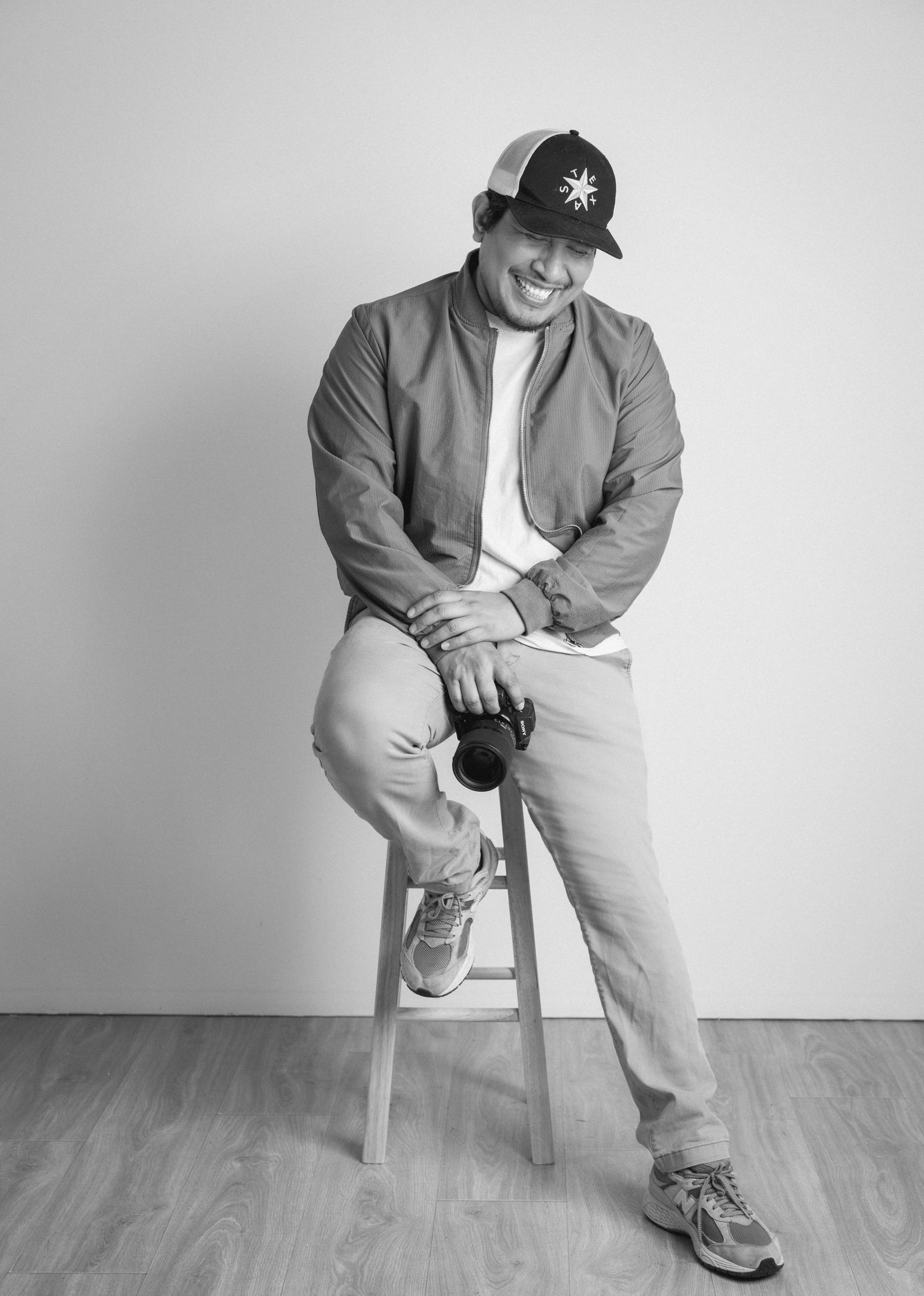 A smiling man in a bomber jacket and cap sits on a stool holding a camera in black and white.