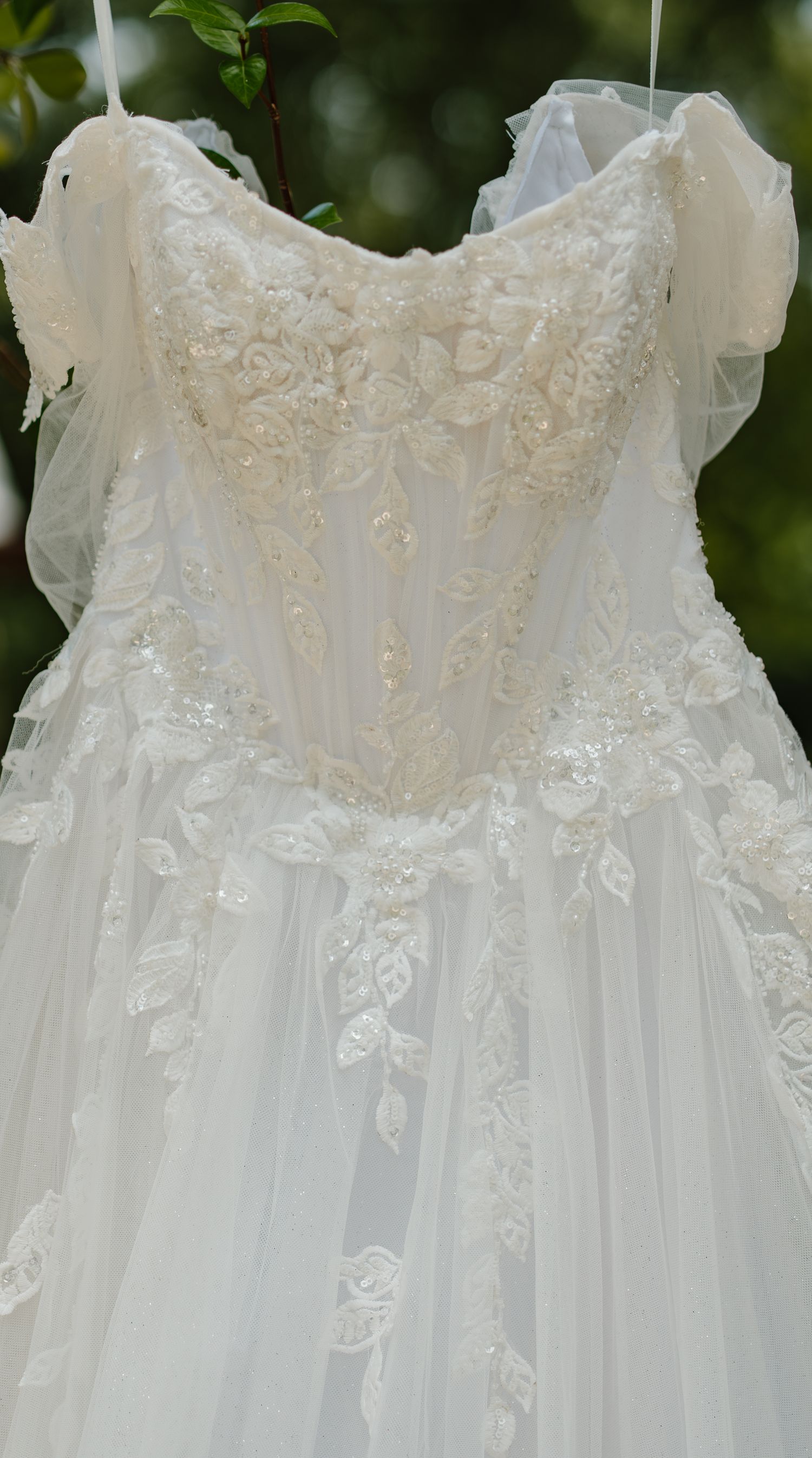 Close-up of an elegant white lace wedding gown with floral embroidery and tulle detailing hanging outdoors.