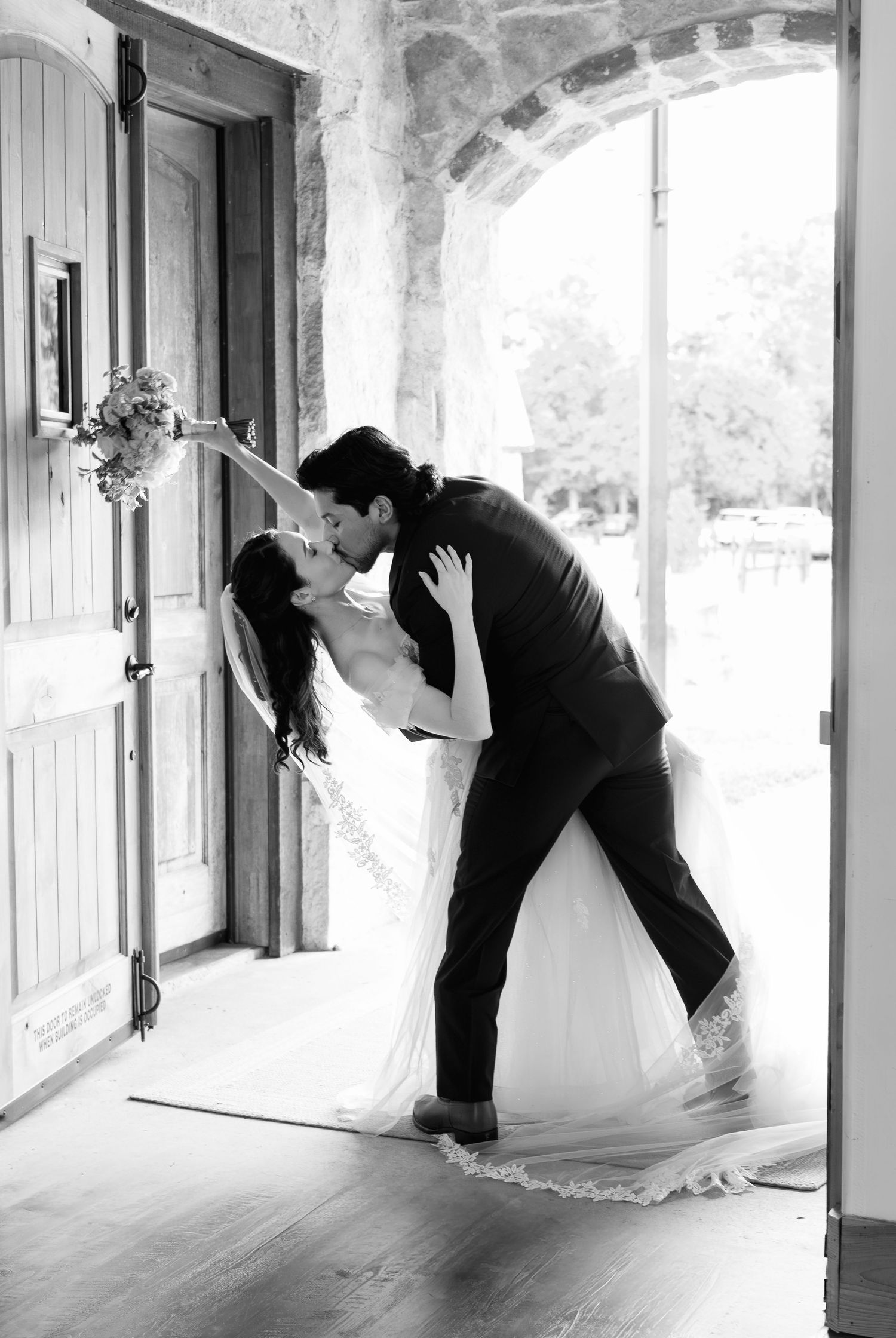 Black and white photo of bride and groom sharing a romantic dip kiss in a rustic stone doorway.