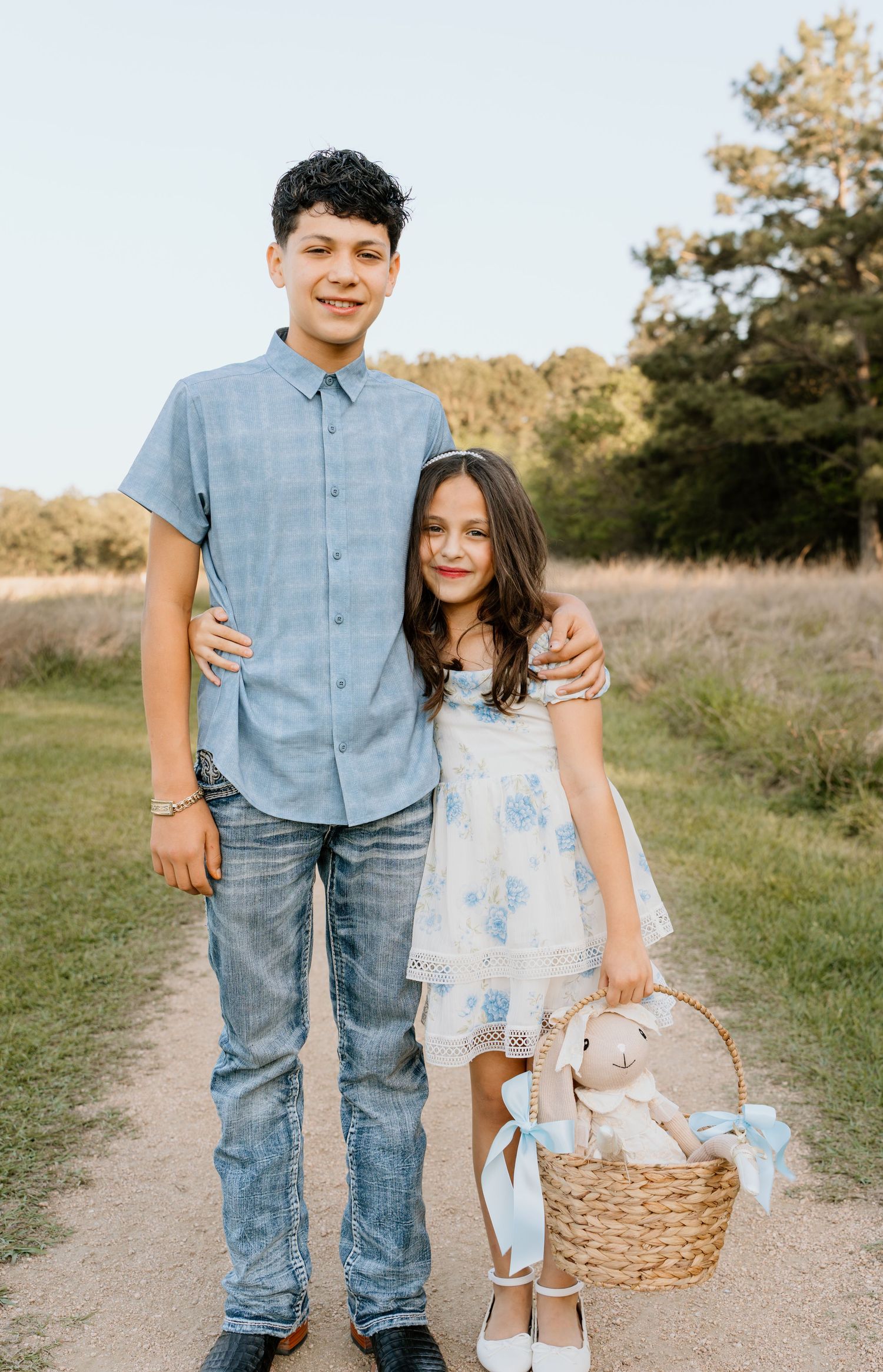 Two children pose on a dirt path, boy in blue shirt and jeans with girl in floral dress holding a wicker basket with stuffed bunny.