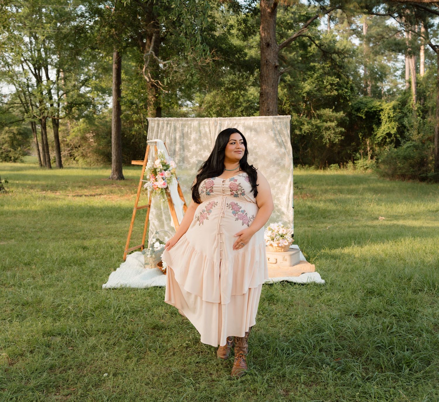 Woman in floral embroidered blush dress poses outdoors at boho garden party setup with lace backdrop.