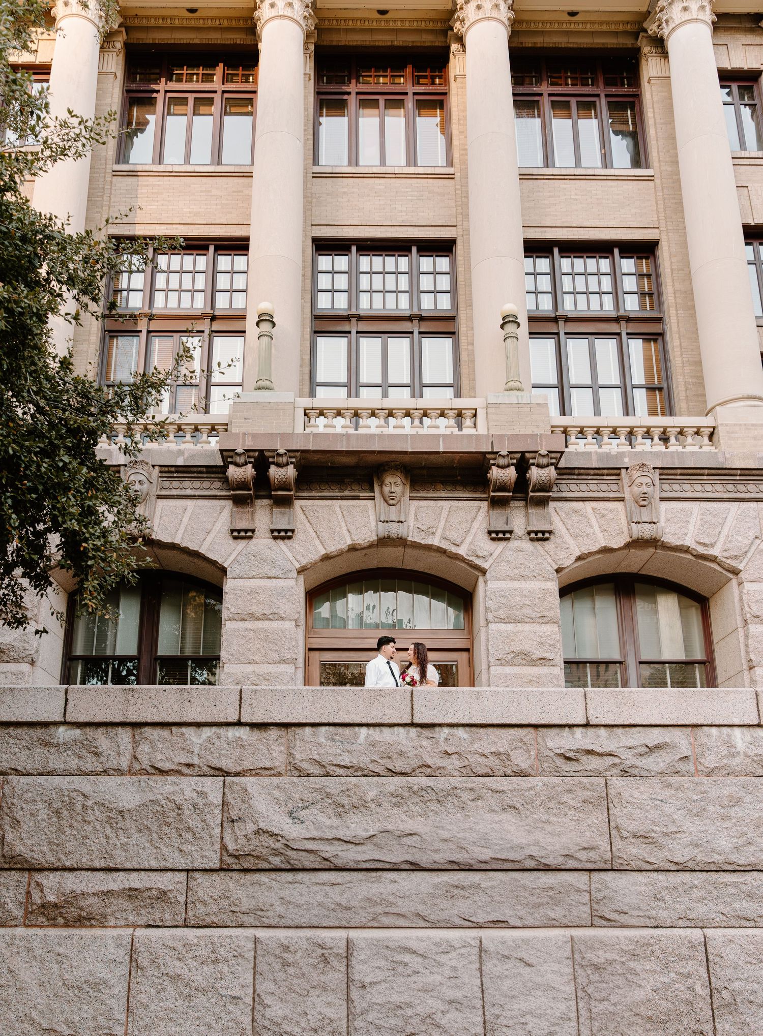Bride and groom share intimate moment on ledge of grand historic stone building with tall windows.