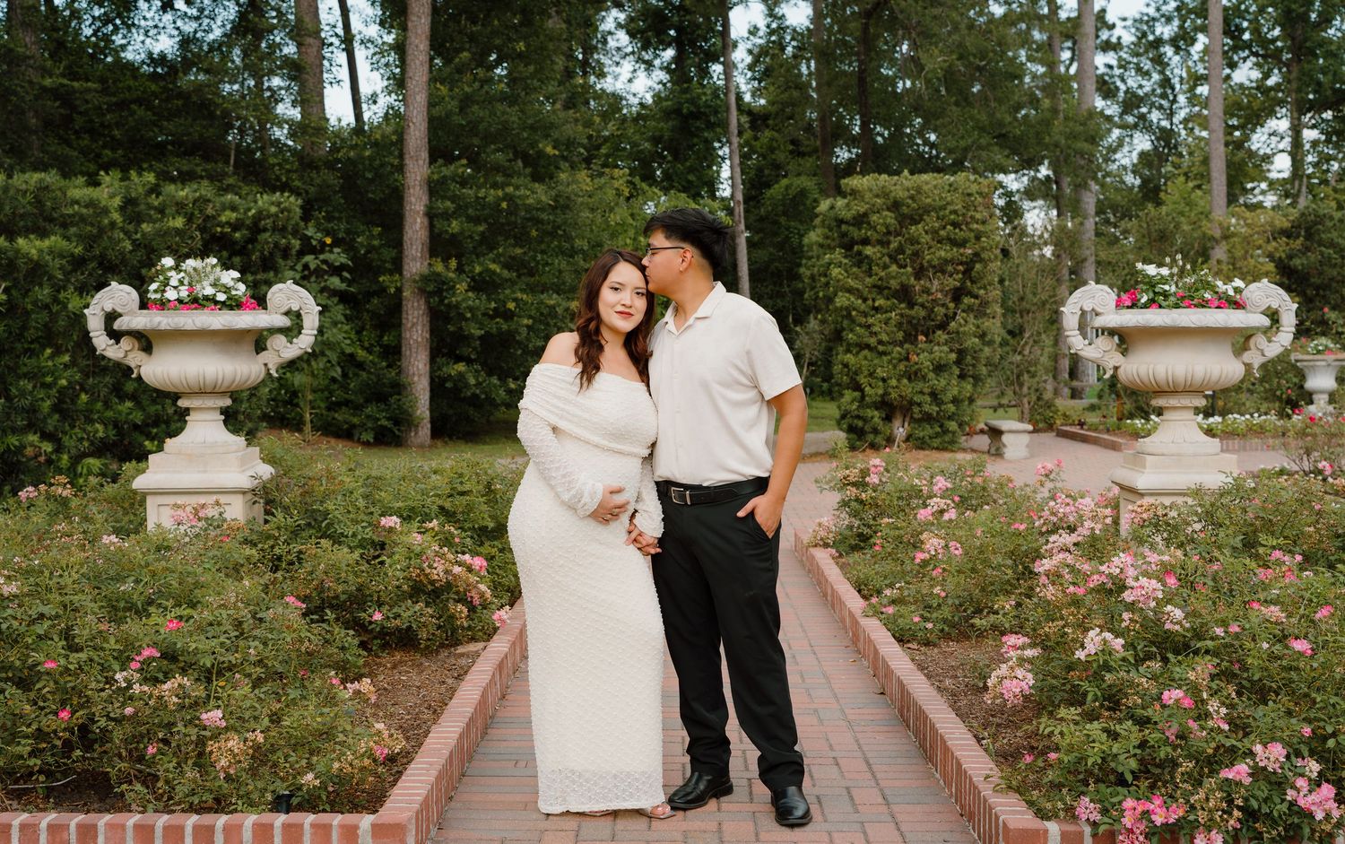 Couple in white dress and black pants sharing a romantic moment in a rose garden with decorative urns.