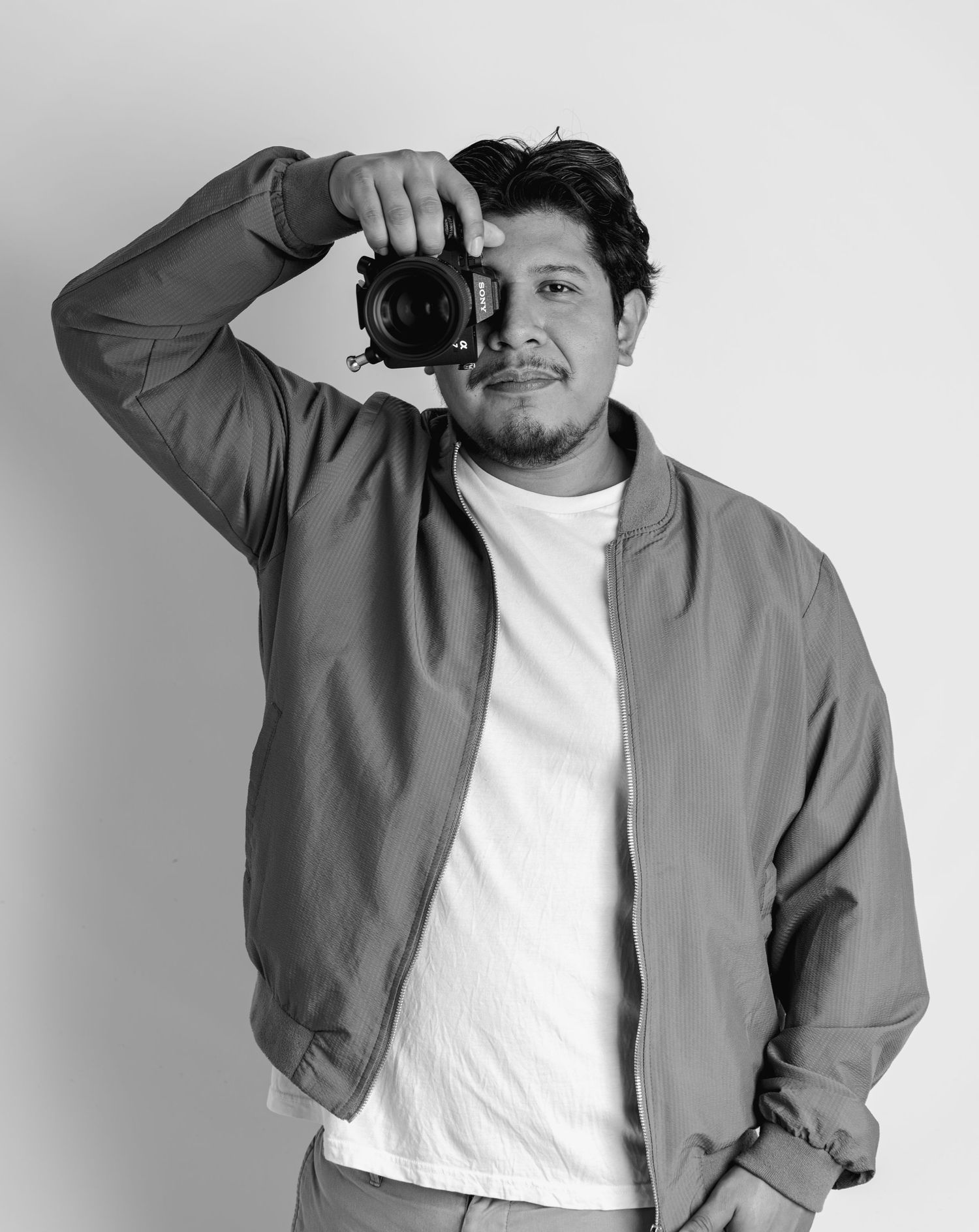 A male photographer raises his camera to shoot in a black and white portrait studio photo.