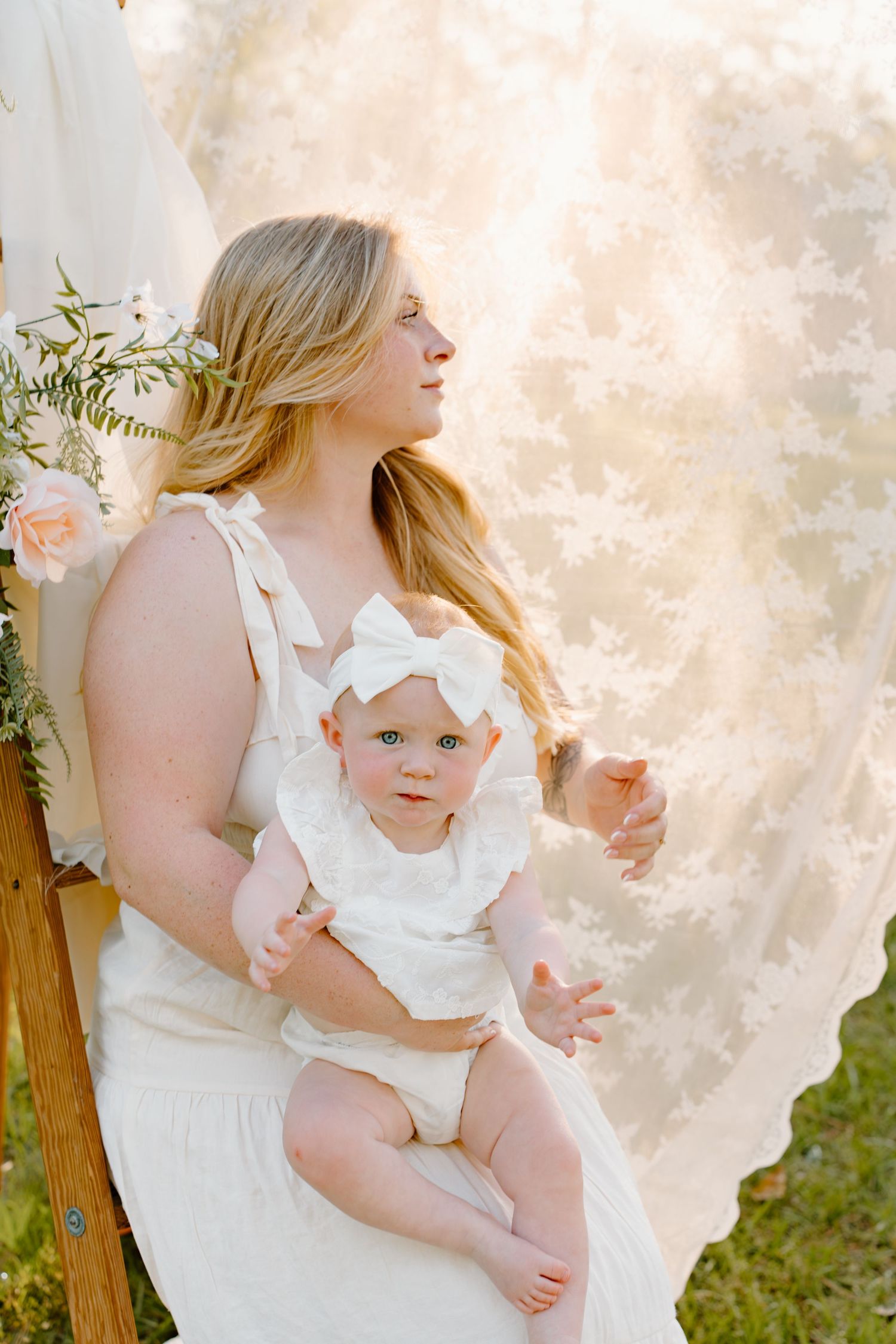 A mother in a white dress holds her baby girl in a white outfit and bow headband during an outdoor photo session.