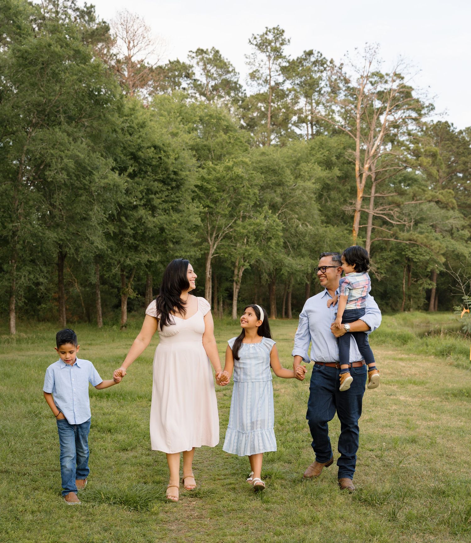 Family of five walking hand-in-hand through a lush green field surrounded by tall trees.