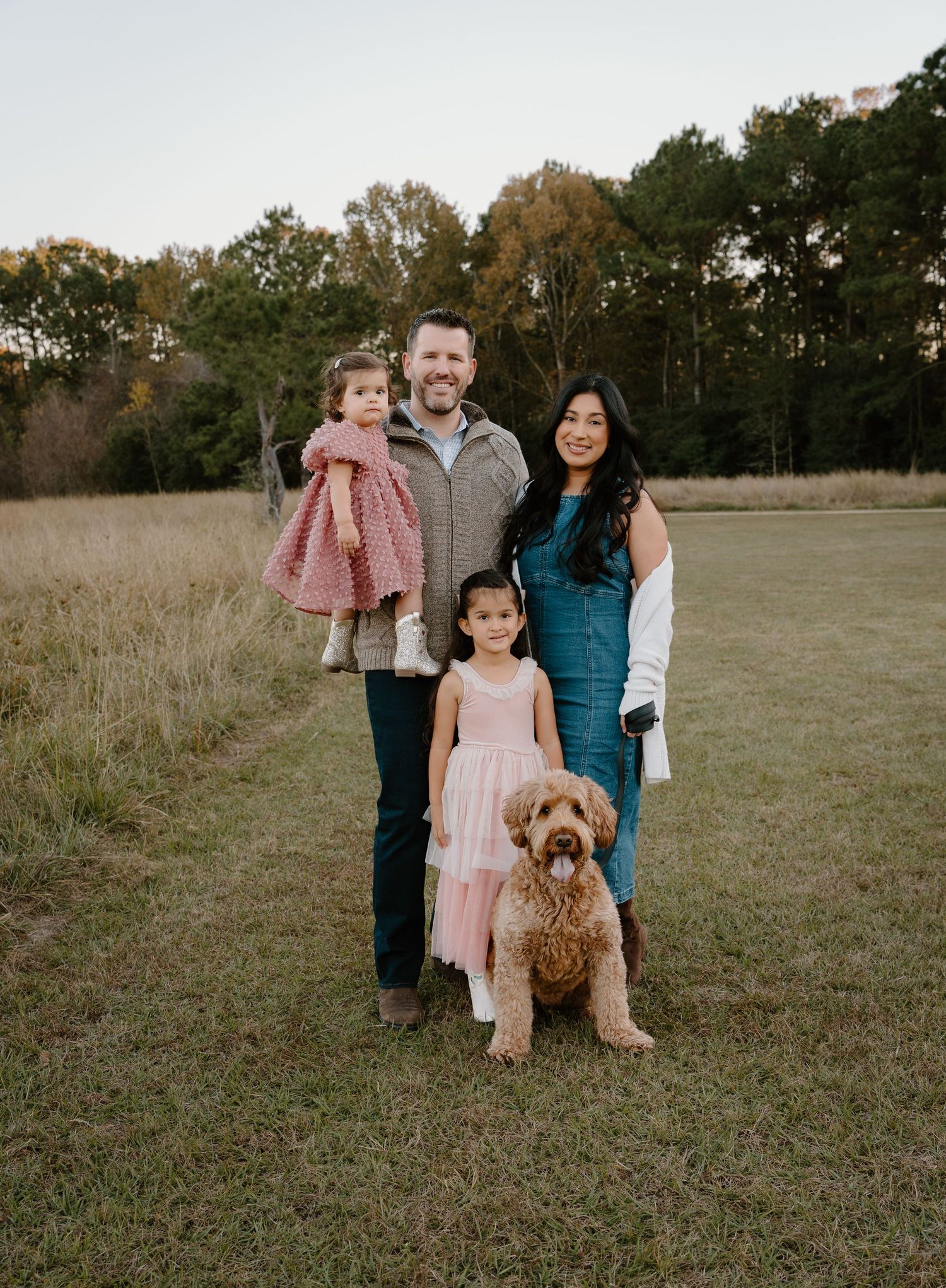 Family of four with a golden doodle dog posing in an open field surrounded by autumn trees.