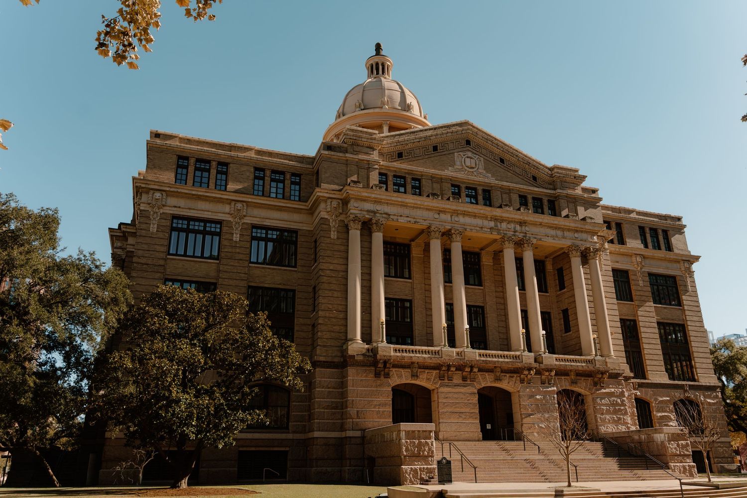 Historic beige stone courthouse with grand columns and dome under clear blue sky surrounded by trees.