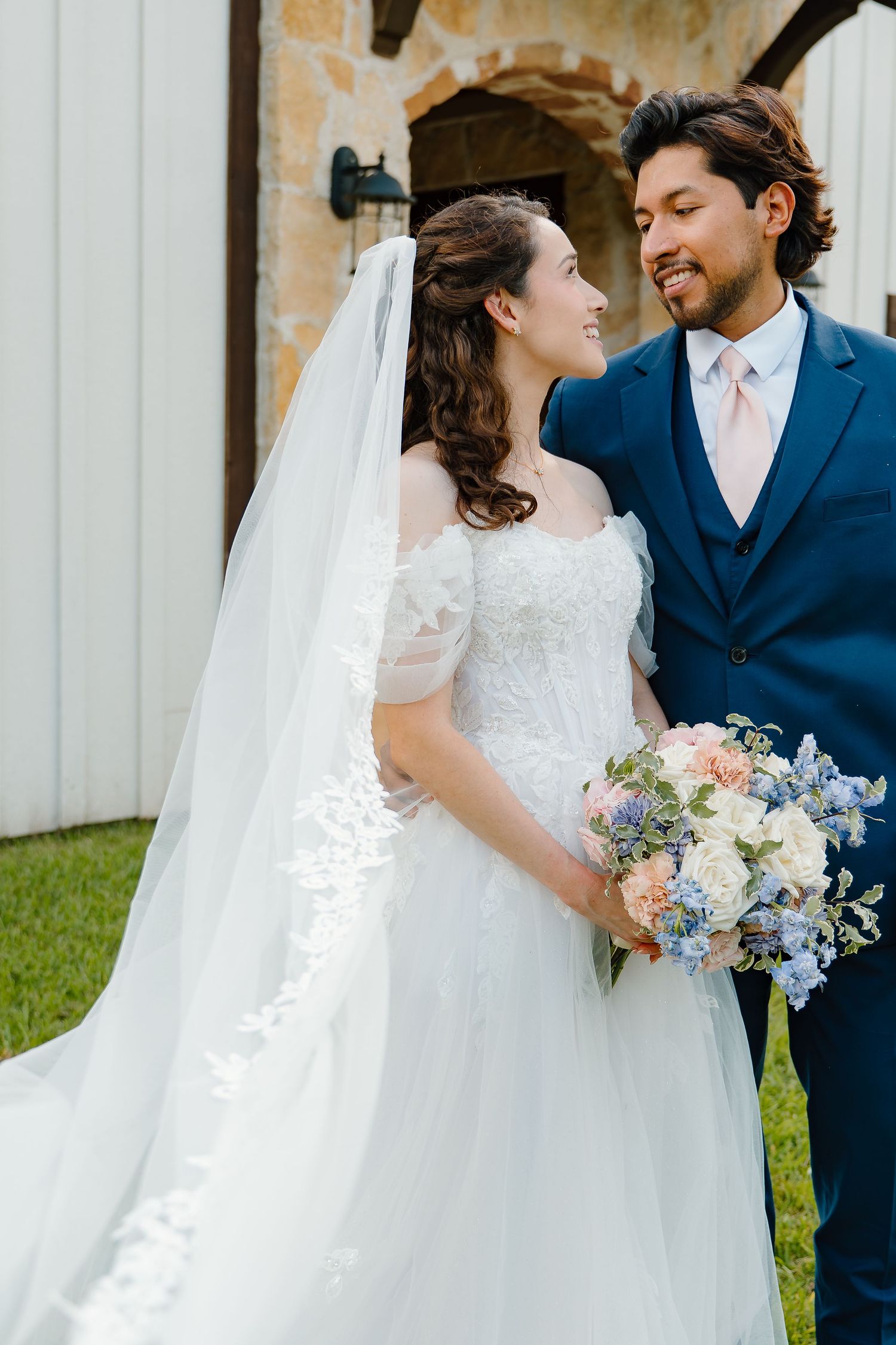 Bride and groom gazing at each other, bride holding pastel bouquet in off-shoulder lace gown with cathedral veil.