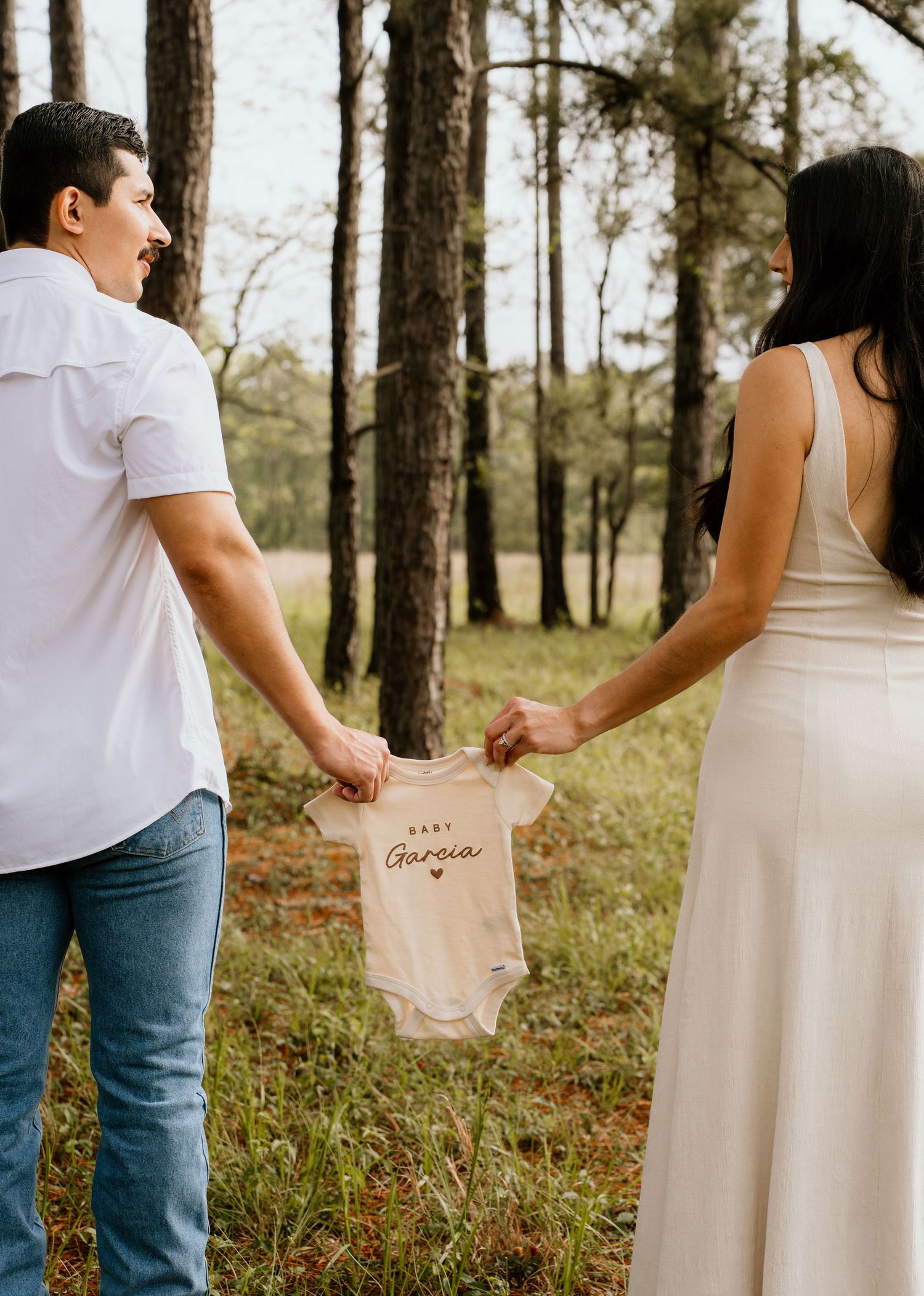 Couple holding a Baby Garcia onesie between them while walking through a pine forest, pregnancy announcement photo.