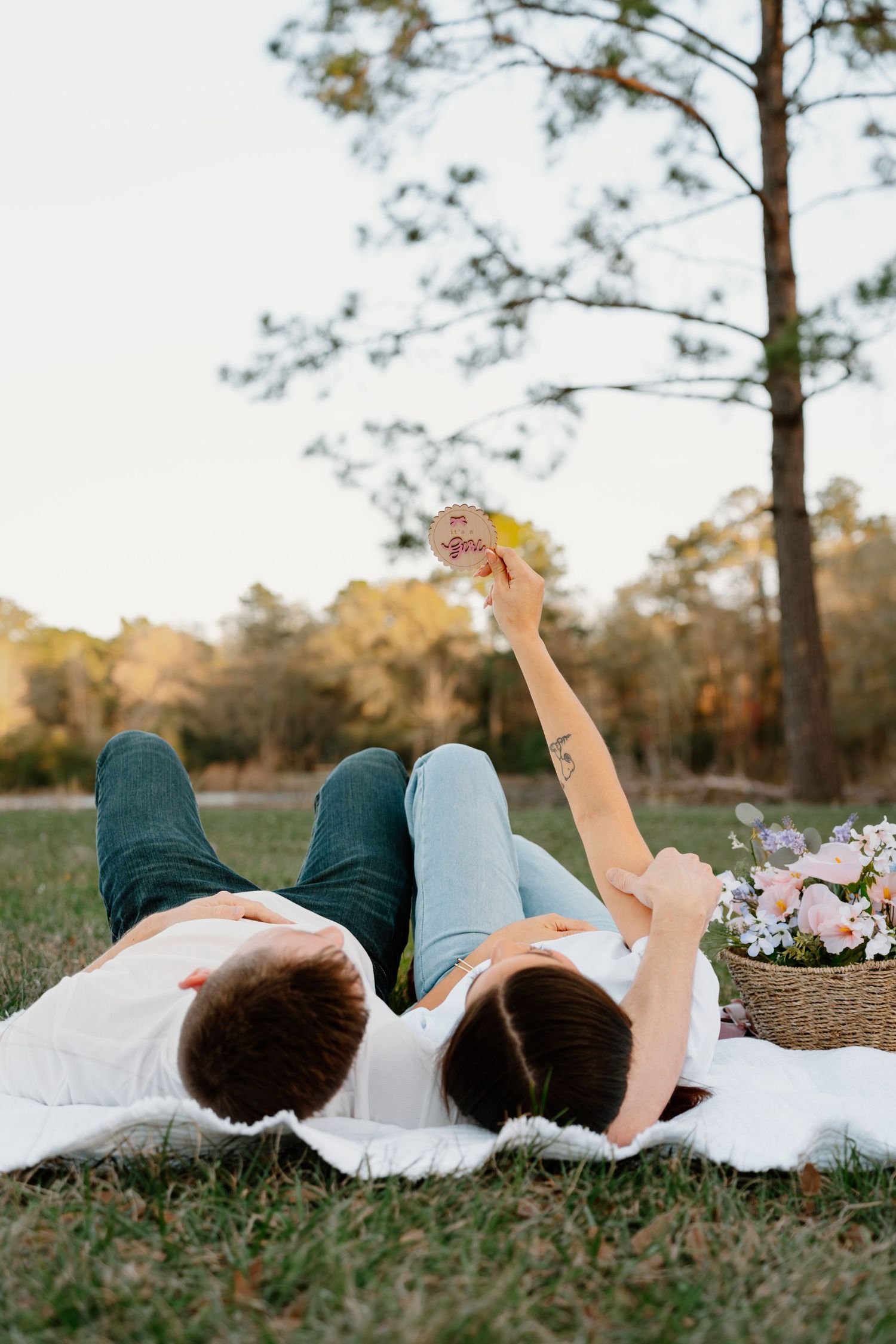 Couple lying on a blanket in a park holding a gender reveal ornament at sunset.