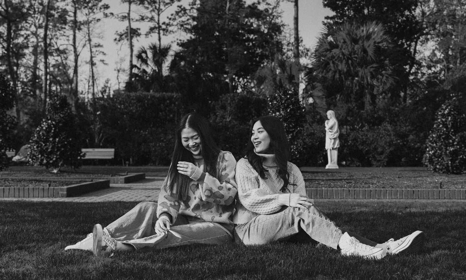 Two young women sit laughing together on grass in a black and white garden portrait photo.