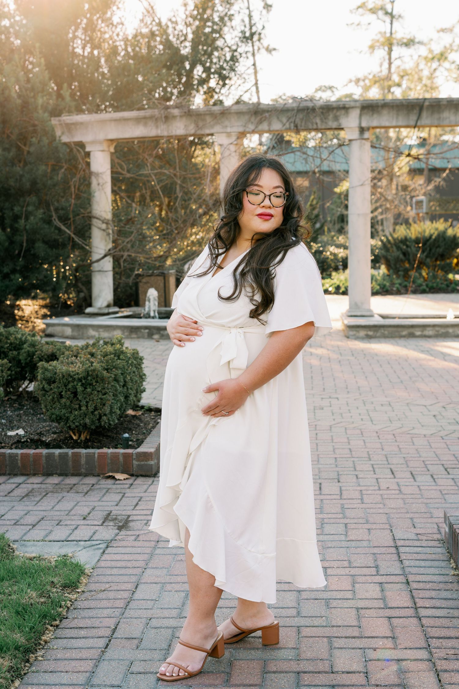 Pregnant woman in white wrap dress and glasses poses on brick path in garden with stone pergola at golden hour.