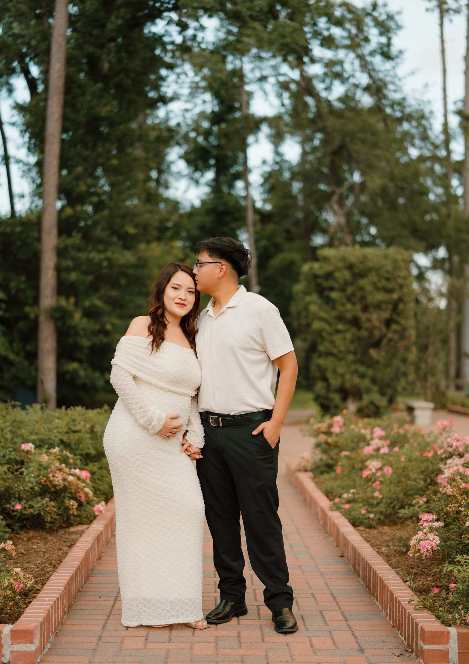 Couple poses in rose garden, woman in white maternity dress, man in polo shirt, brick pathway, lush greenery backdrop.