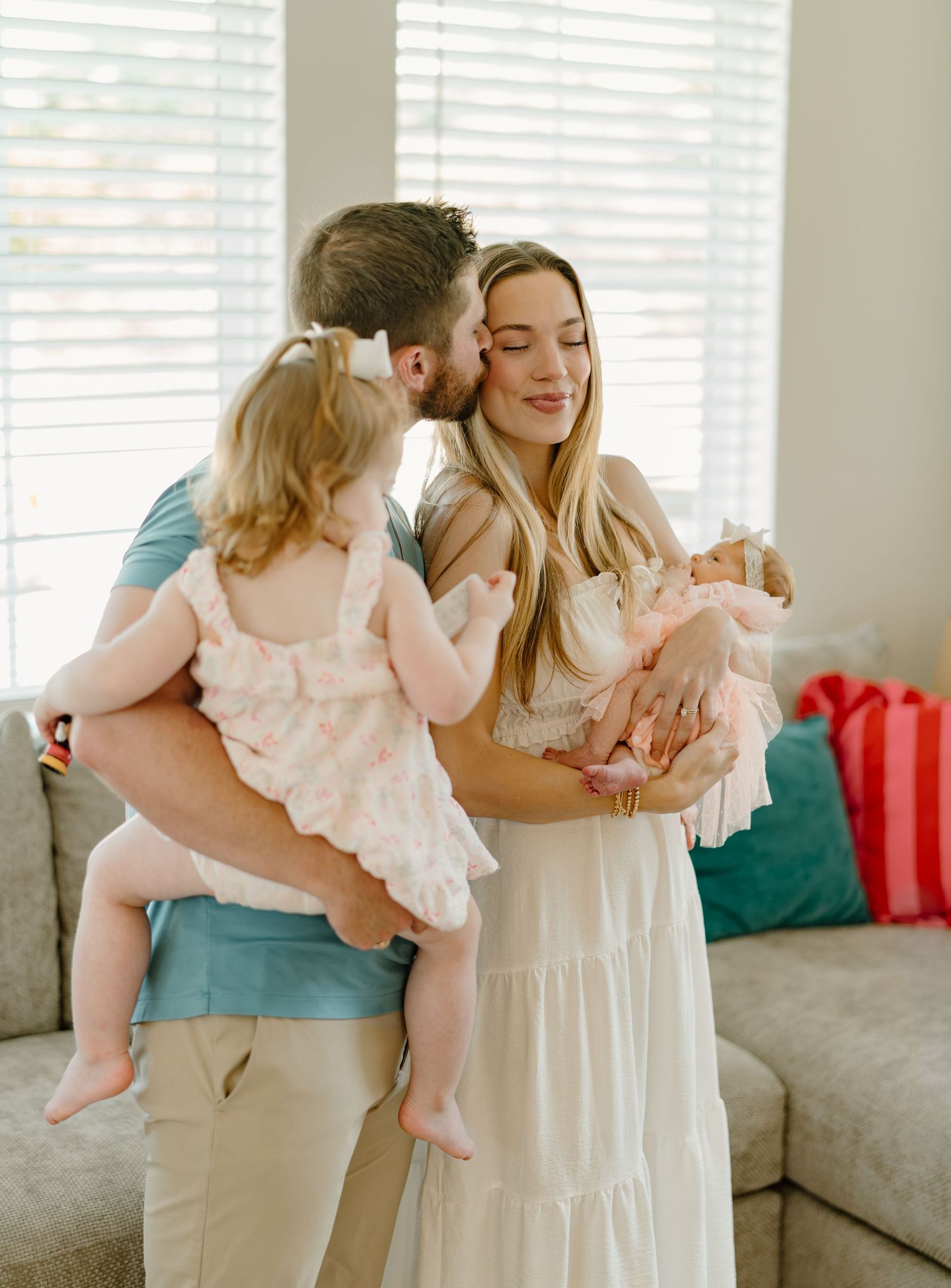 A couple shares a tender kiss while holding their toddler and newborn baby in a bright, cozy living room.
