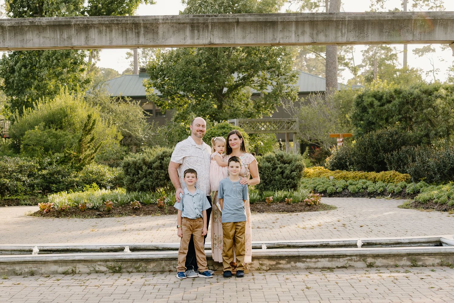 Family of four posing in a lush garden park, parents with two young boys wearing matching tan pants.