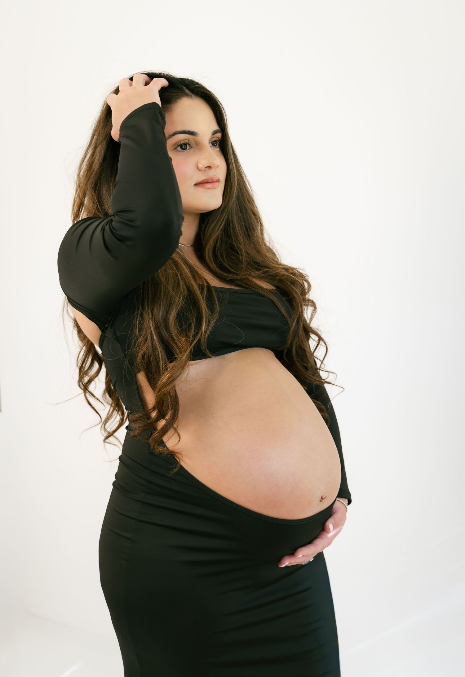 Pregnant woman in black crop top and skirt cradling baby bump, with long brunette hair, posing against white background.