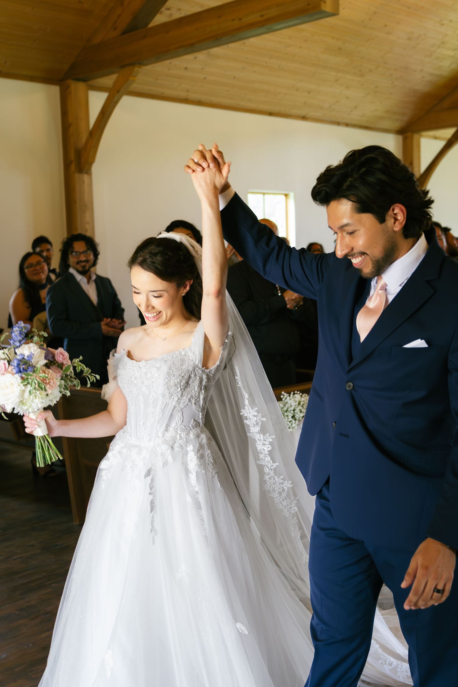 Joyful newlyweds raise hands in celebration walking down the aisle, bride in lace gown holding bouquet.