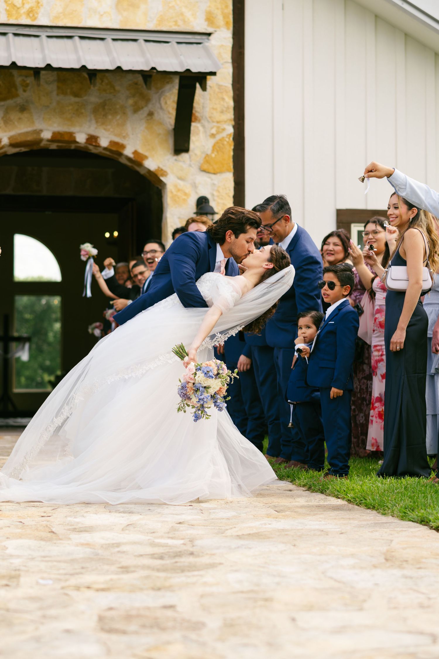 Bride and groom share a romantic dip kiss outside a rustic stone barn surrounded by cheering wedding guests.