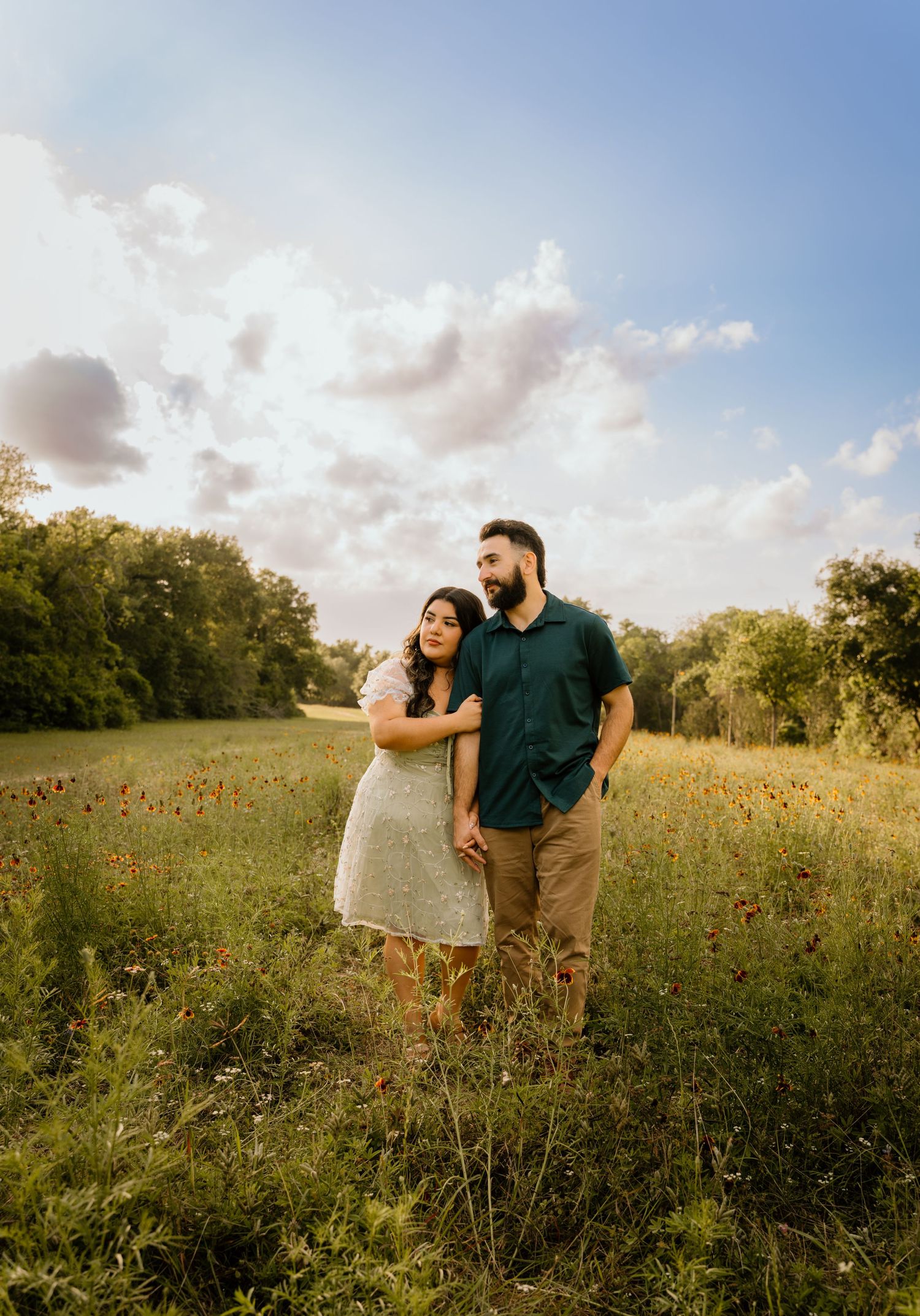 Couple embraces while holding hands in colorful wildflower field beneath beautiful blue cloudy sky at sunset.